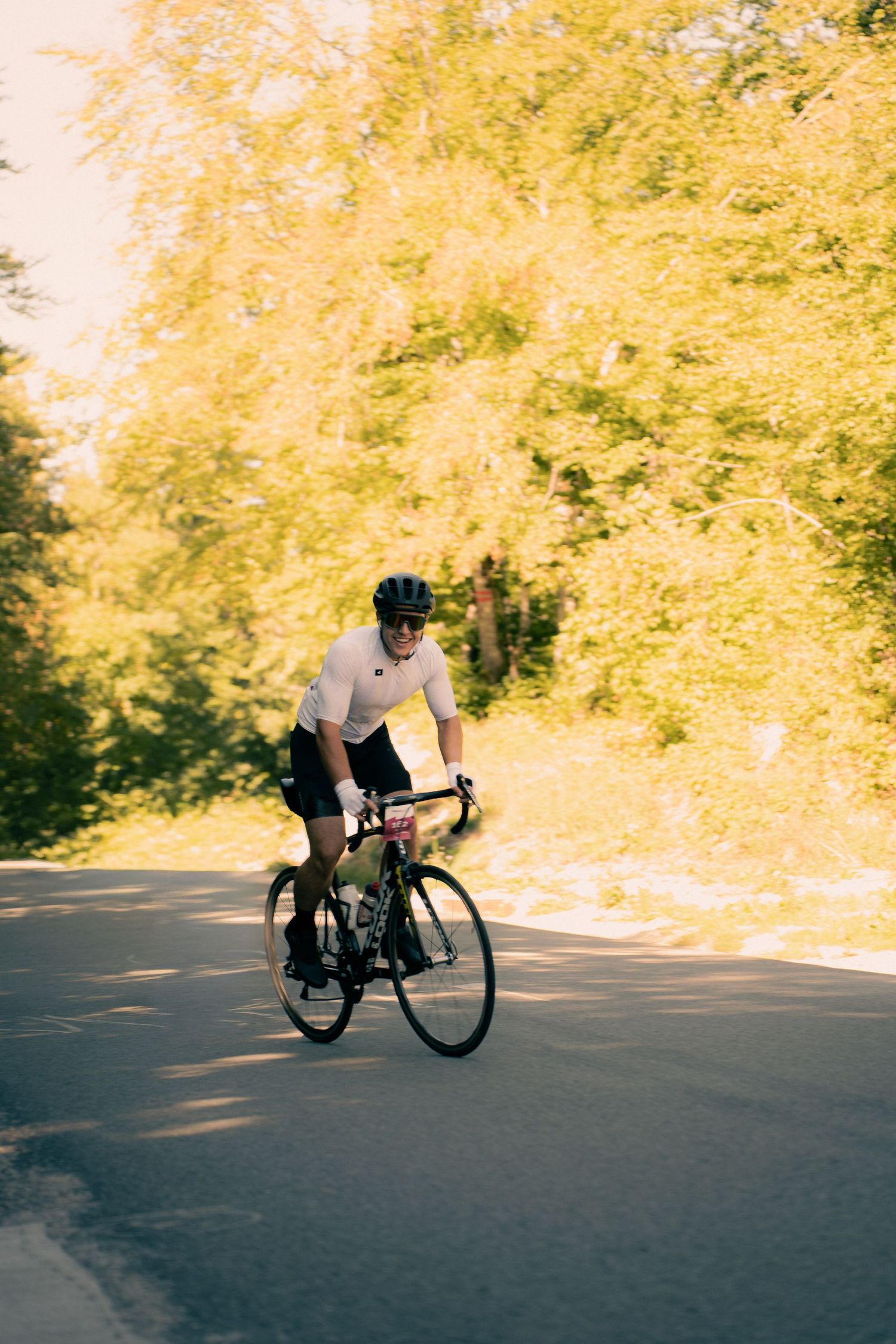 Cyclist in white jersey and helmet riding a road bike on a sunny tree-lined path.
