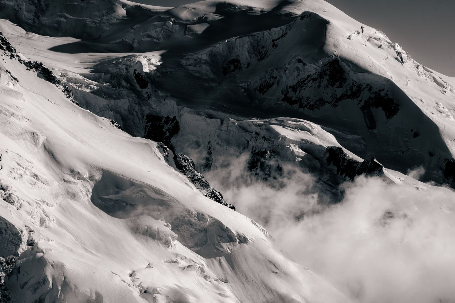 Black and white aerial view of snow-covered mountain peaks with dramatic shadows and swirling clouds below.