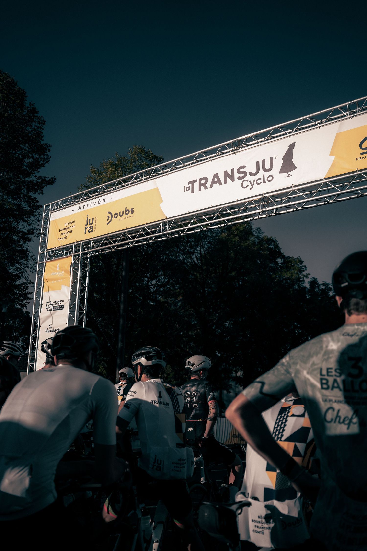 Cyclists gather at the start line of la TransJu Cyclo race under a branded arch in a dramatic outdoor setting.