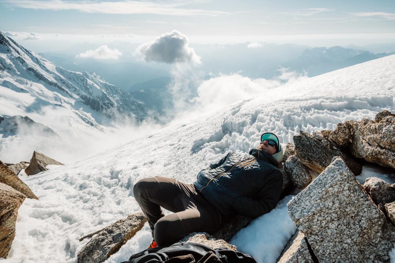 Mountain climber rests on rocky ledge with stunning snowy glacier and cloud-filled valley below.