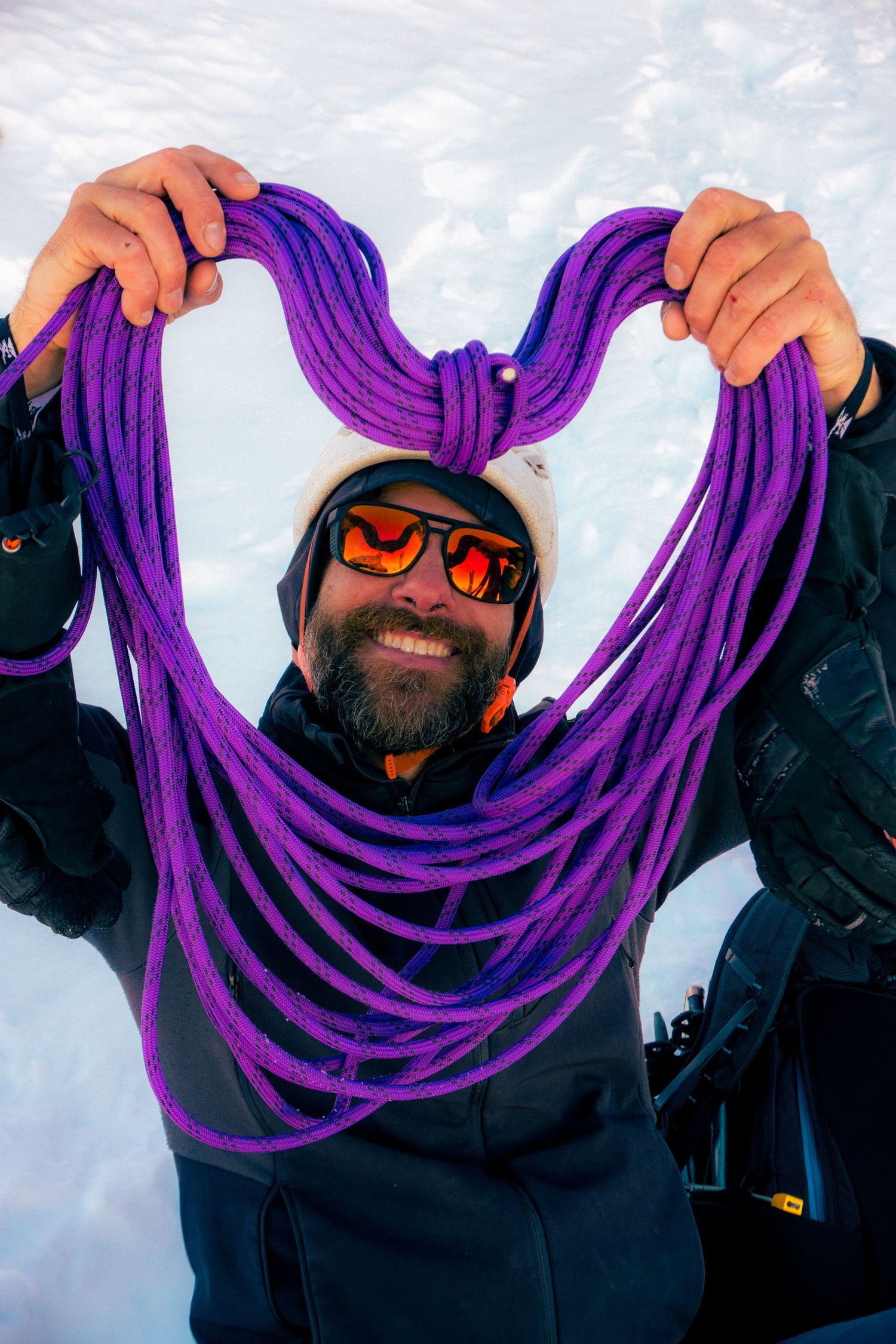 Smiling bearded man in snowy mountains holding purple climbing rope in heart shape, wearing orange reflective sunglasses.