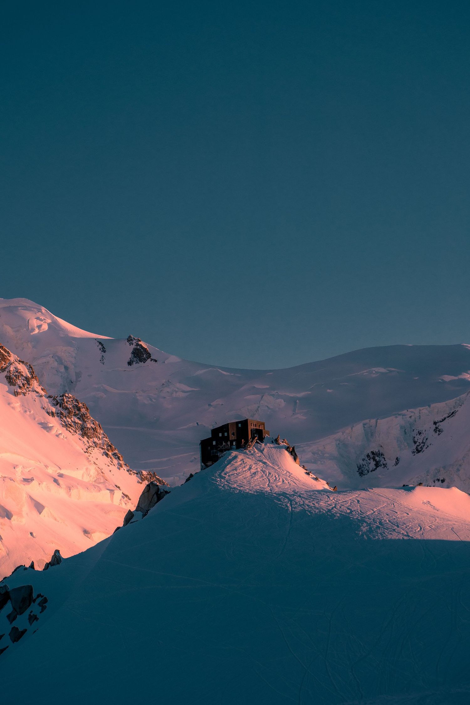 A mountain refuge hut perched on a snowy ridge at sunset with Mont Blanc massif glowing orange behind.