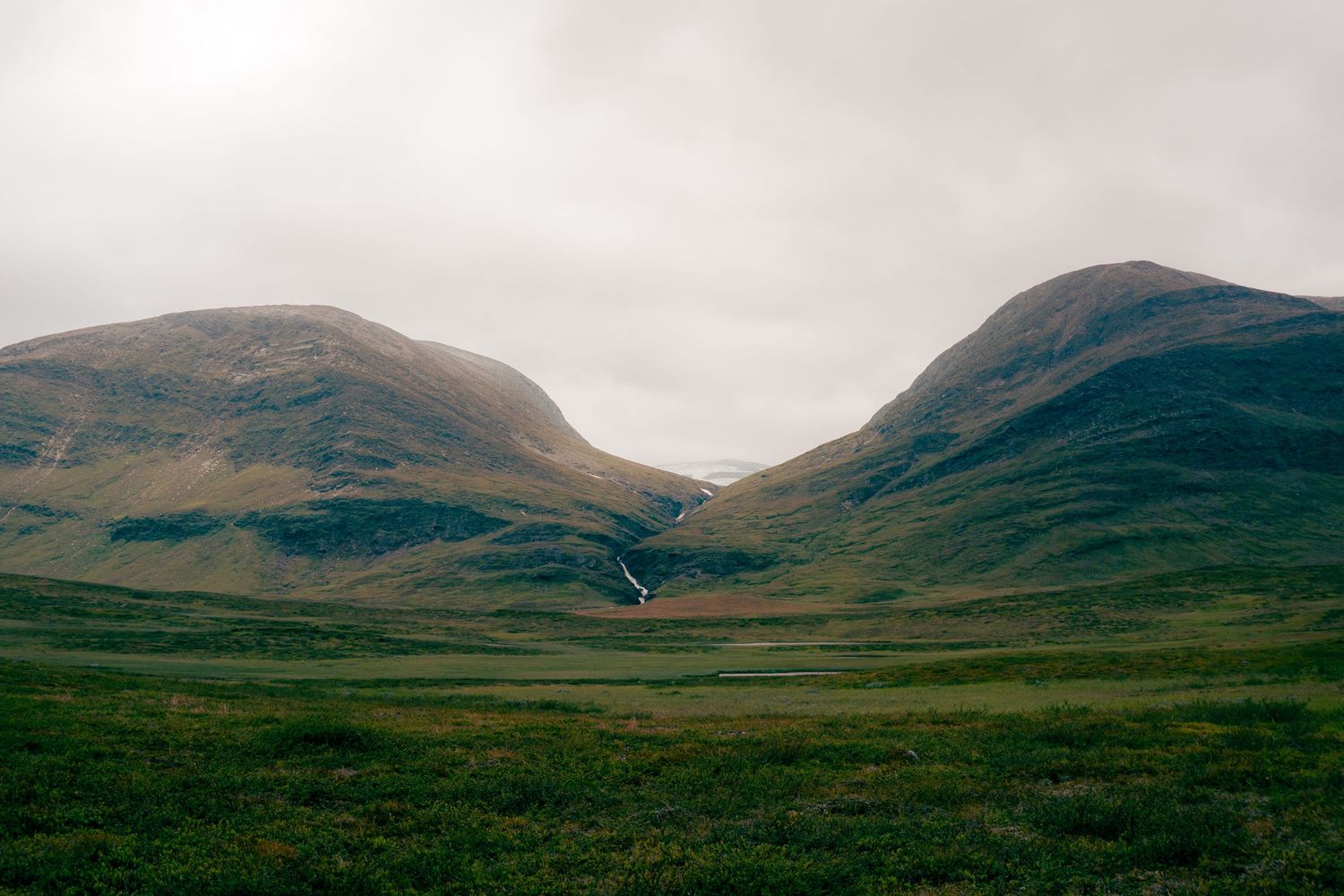 Two rounded green mountains with a small waterfall between them under a cloudy grey sky in Scandinavia.