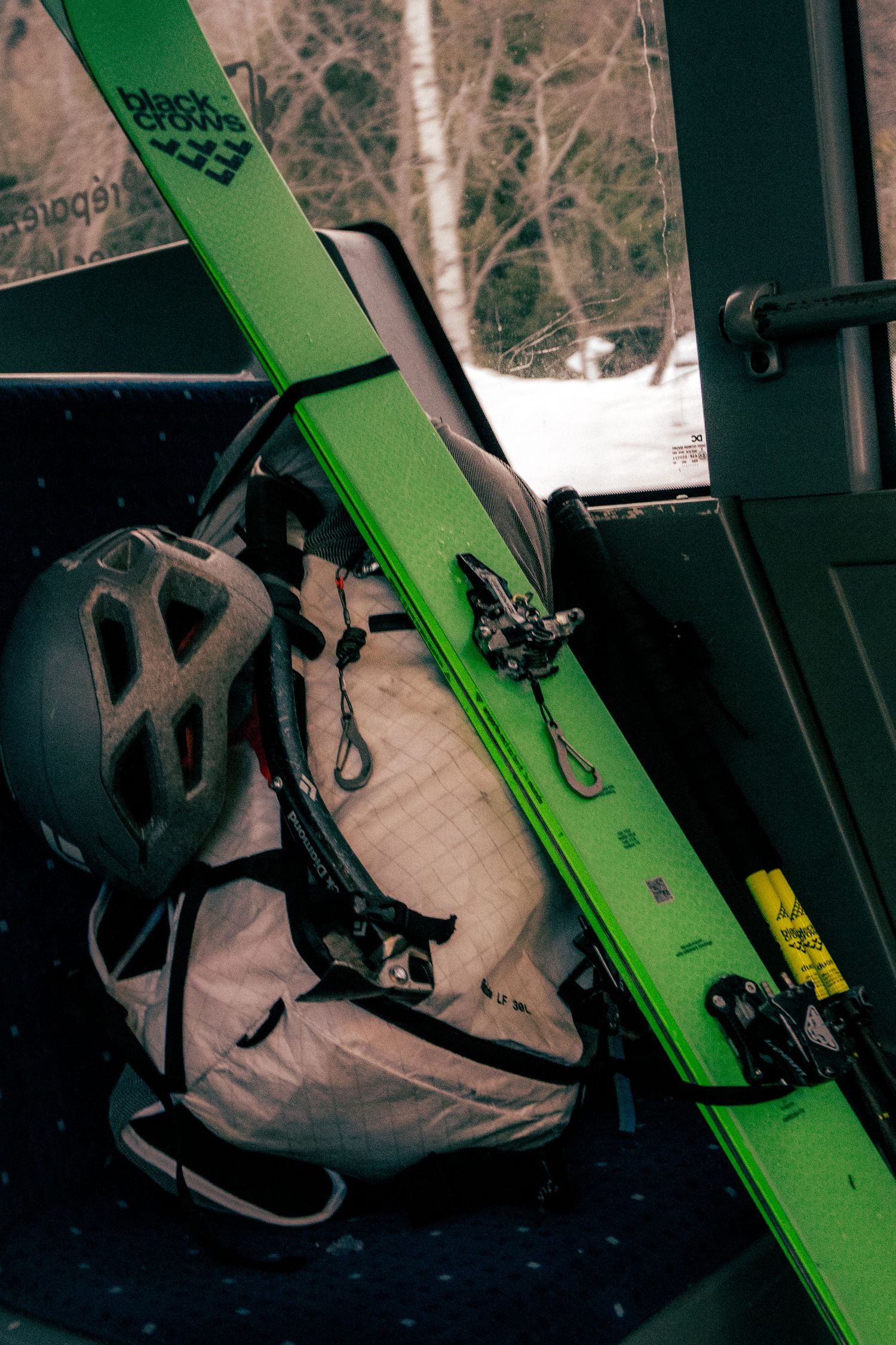 Green Black Crows skis leaning against a gondola wall next to a gray helmet and backpack at a ski resort.