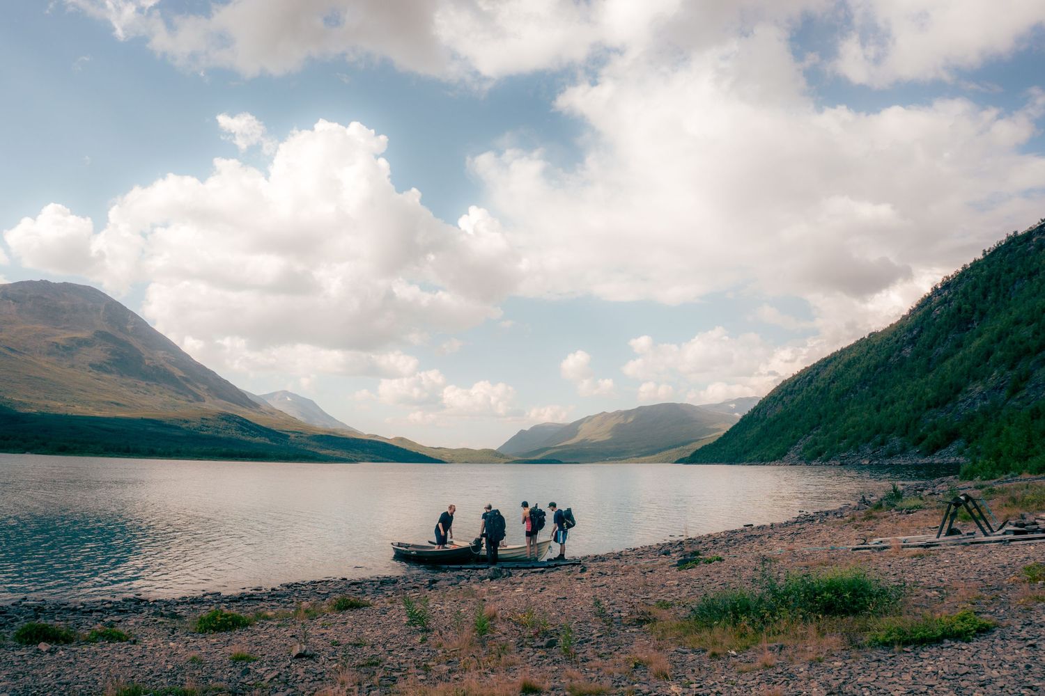 Group of hikers standing by a serene mountain lake with rocky shoreline and dramatic cloudy sky.