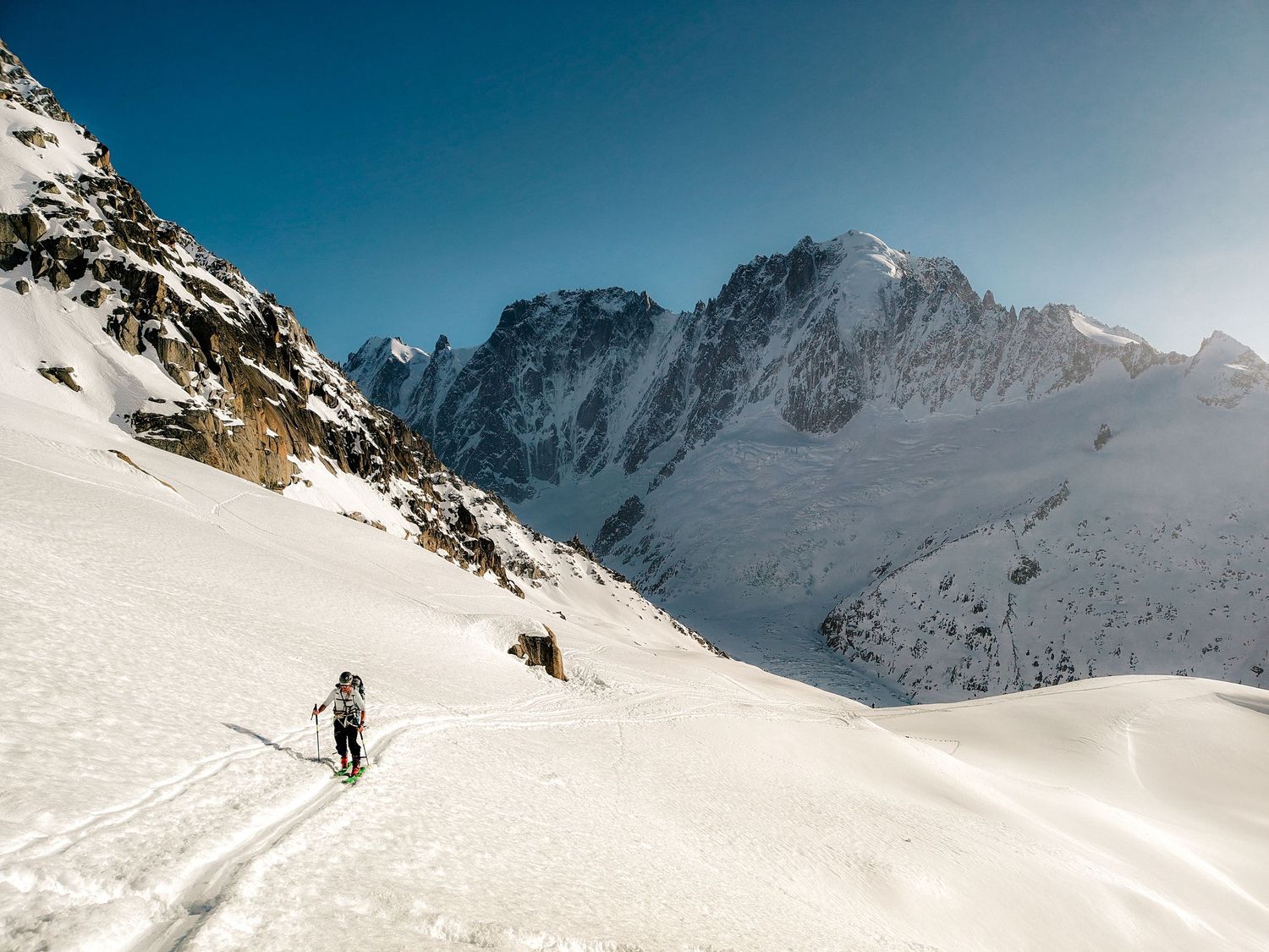 A lone skier ascends a vast snowy mountain slope under a clear blue sky with rugged alpine peaks in the background.