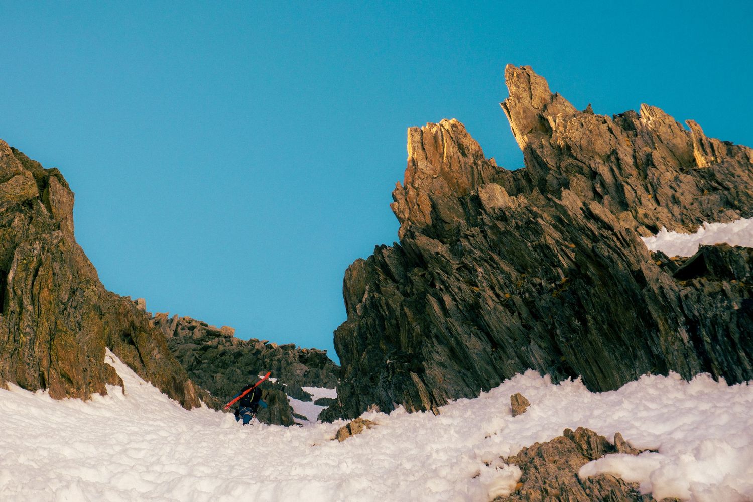A lone climber ascends a snowy mountain slope surrounded by dramatic rocky spires under a clear blue sky.