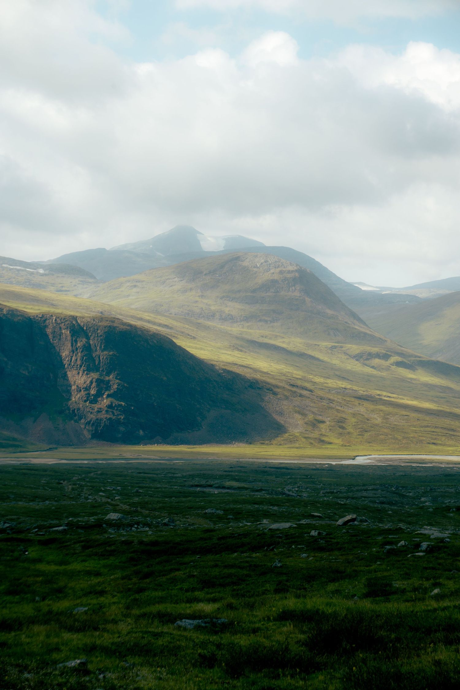 Lush green valley with steep rocky cliffs and rolling mountains partially covered by low clouds.