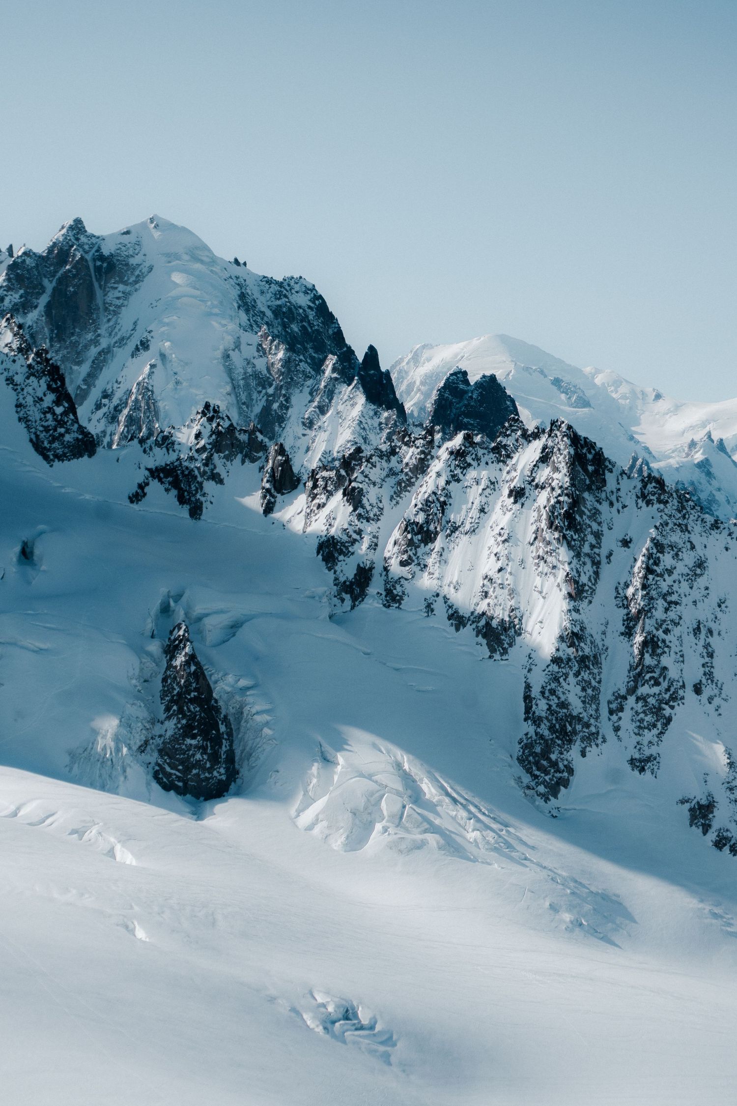 Snow-covered alpine peaks with dramatic rocky ridges rise against a clear pale blue sky in a stunning winter mountain landscape.