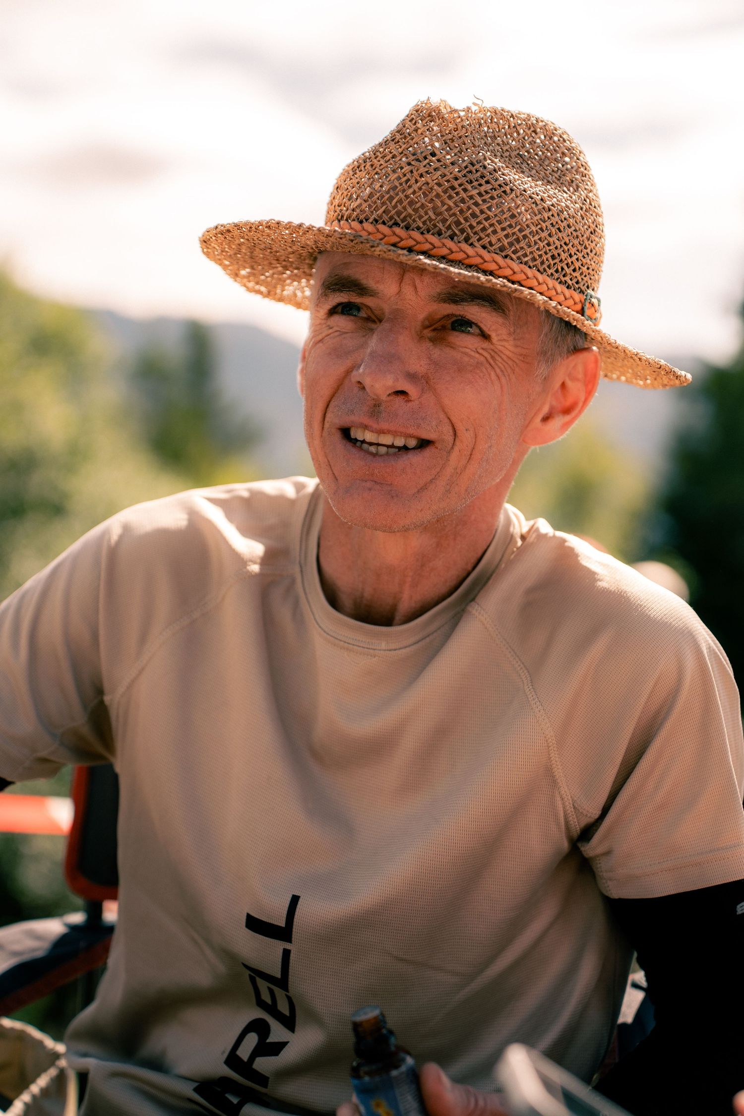 Smiling older man wearing straw hat and Merrell t-shirt resting outdoors in mountain setting.