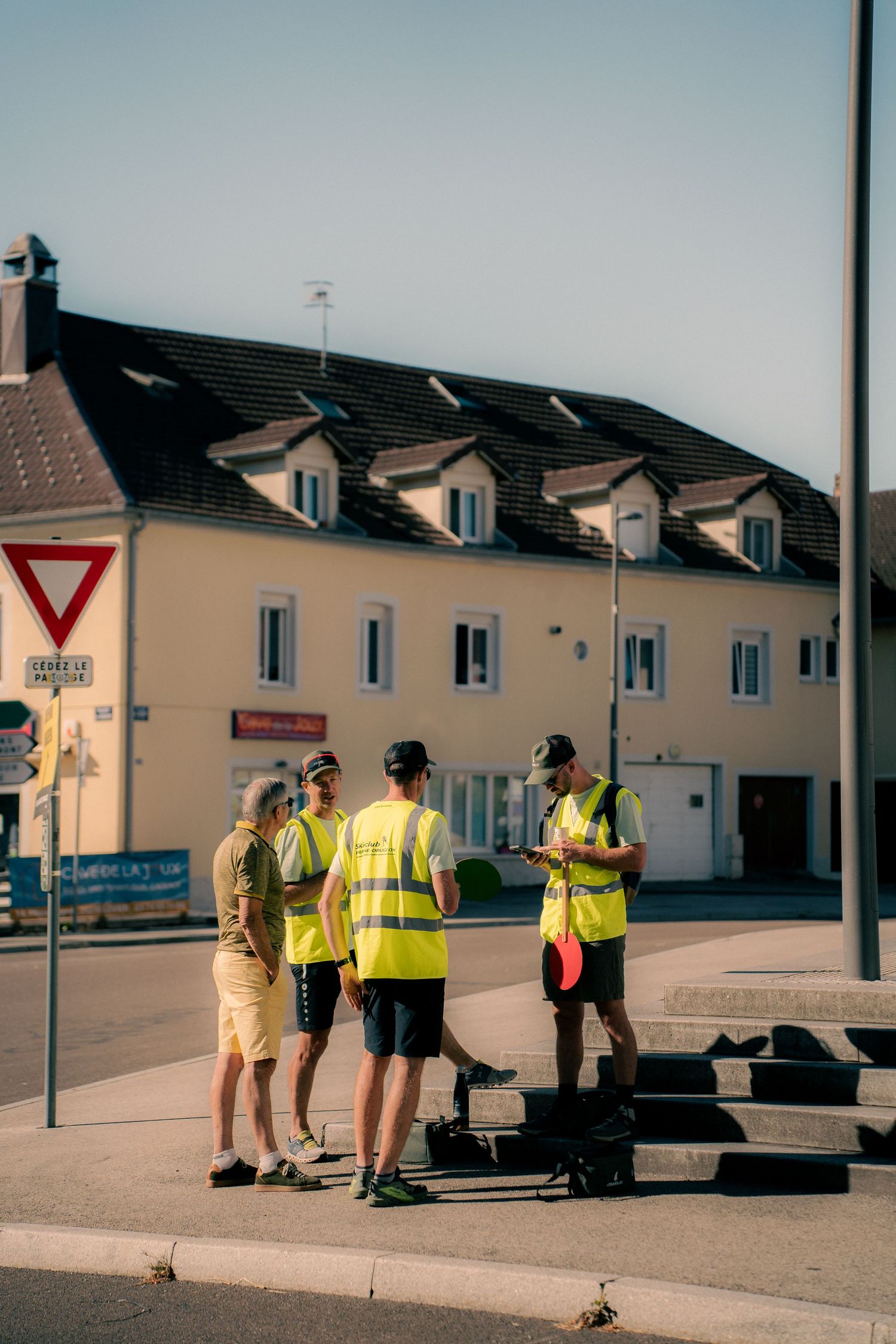 Workers in high-visibility vests discuss plans at a street corner near a yellow building with a yield sign.