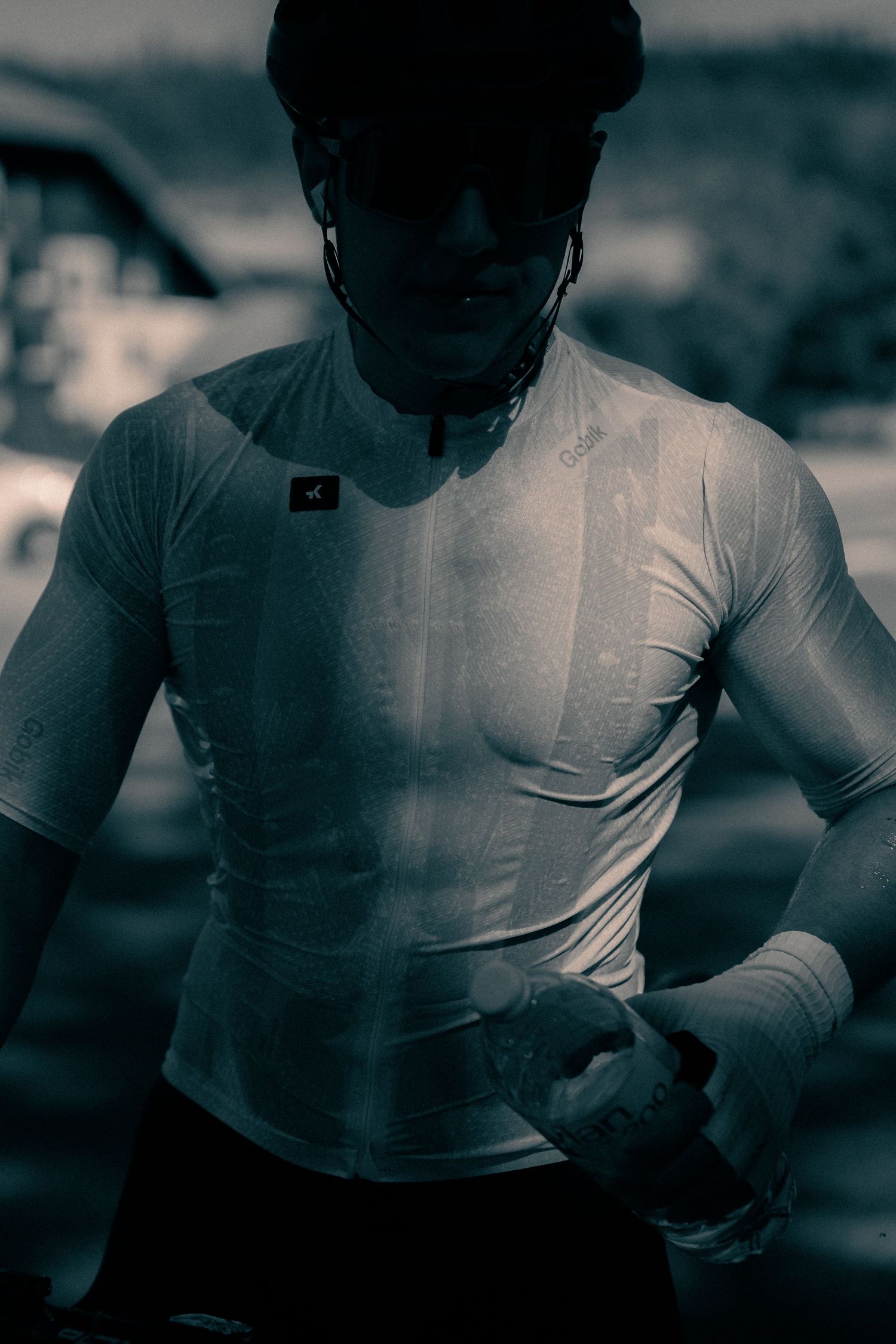 A muscular cyclist in a white long-sleeve jersey and helmet stands outdoors in a dramatic black and white photo.