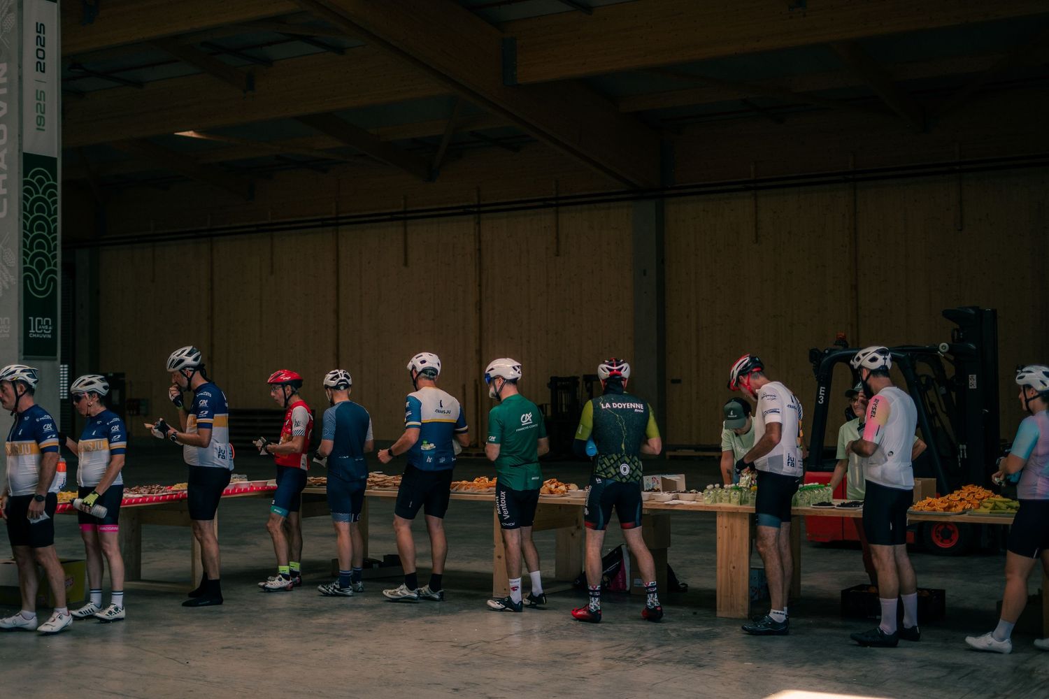 Cyclists in helmets line up at a fruit and food station inside a large warehouse during a cycling event.