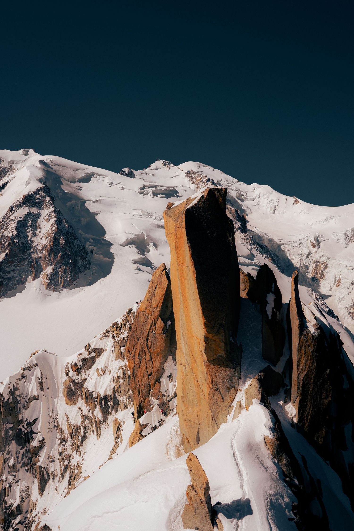 Snow-covered mountain peaks with dramatic rocky spires bathed in golden sunlight against a clear deep blue sky.