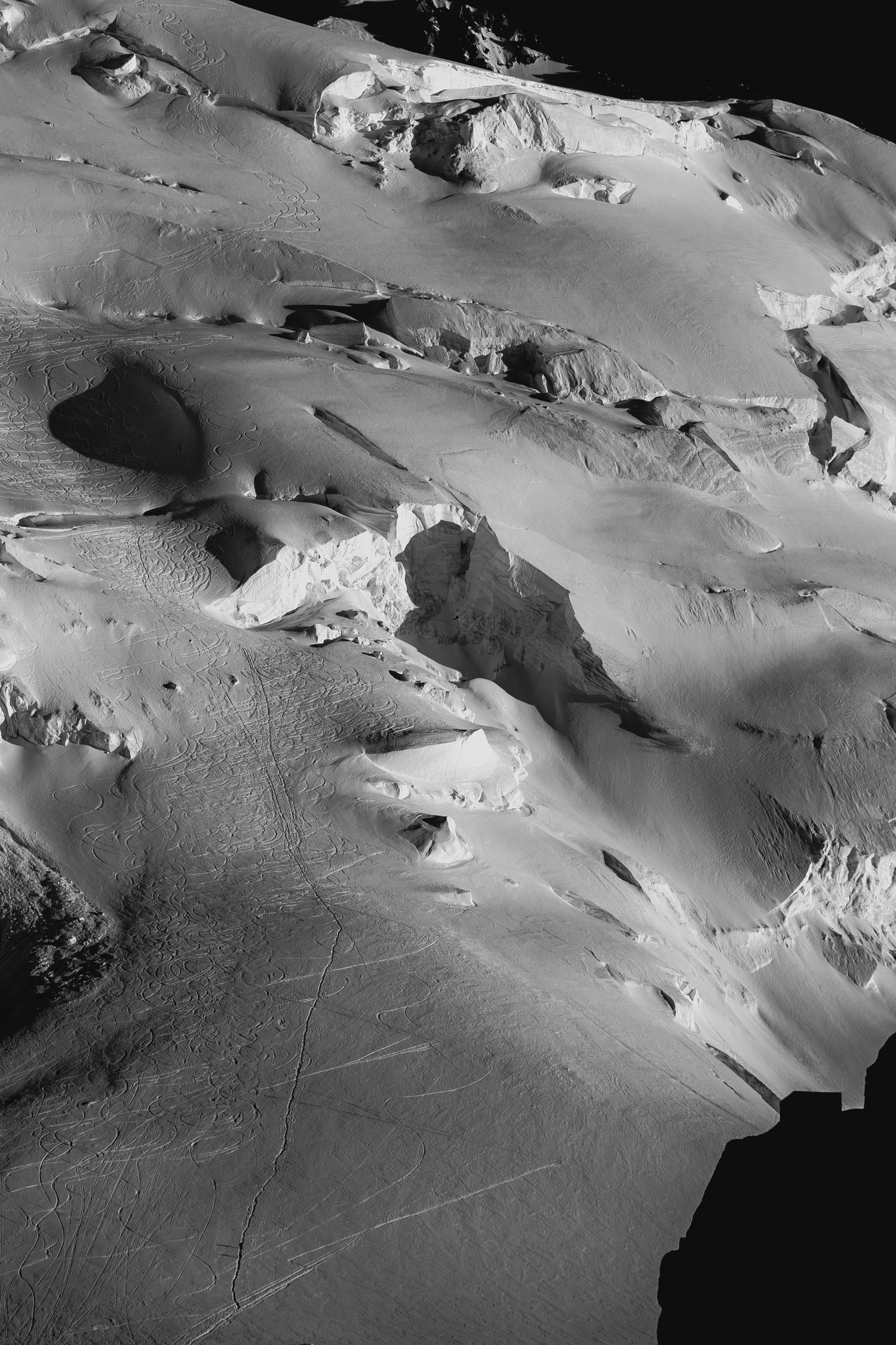 Aerial black and white photo of a snow-covered mountain slope with dramatic shadows and rocky outcroppings.