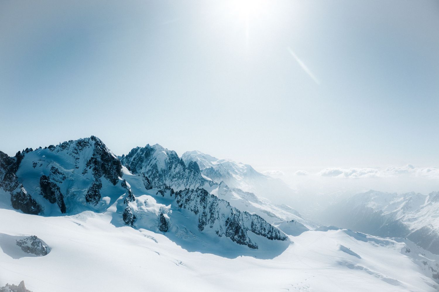 Snow-covered mountain peaks under a bright sun with misty valleys and clear blue sky in a stunning alpine winter landscape.