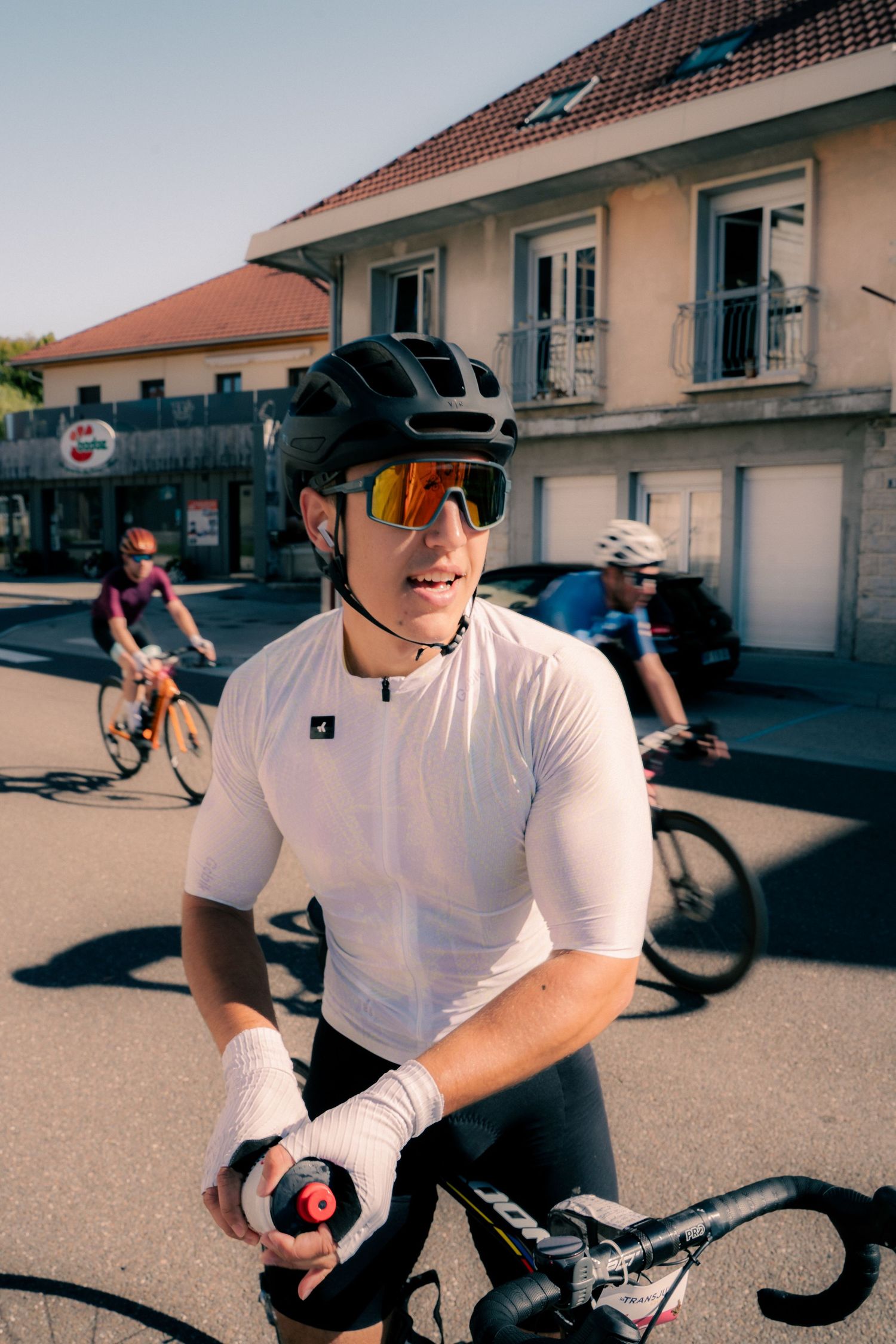 A cyclist in a white jersey, black helmet, and mirrored sunglasses pauses on a sunny road with other riders behind him.