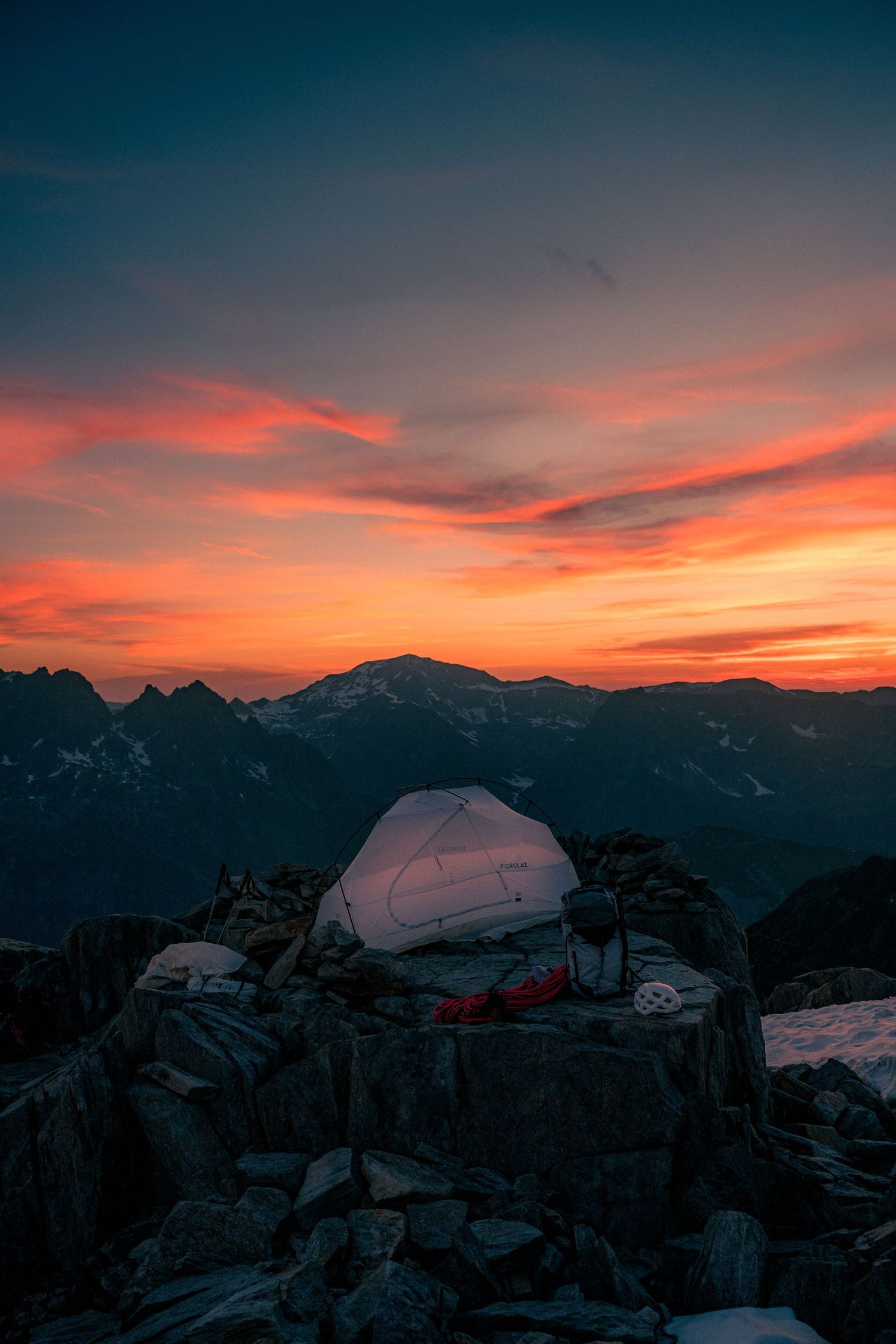 Glowing tent on rocky mountain summit during dramatic red and orange sunset with snow-capped peaks in background.
