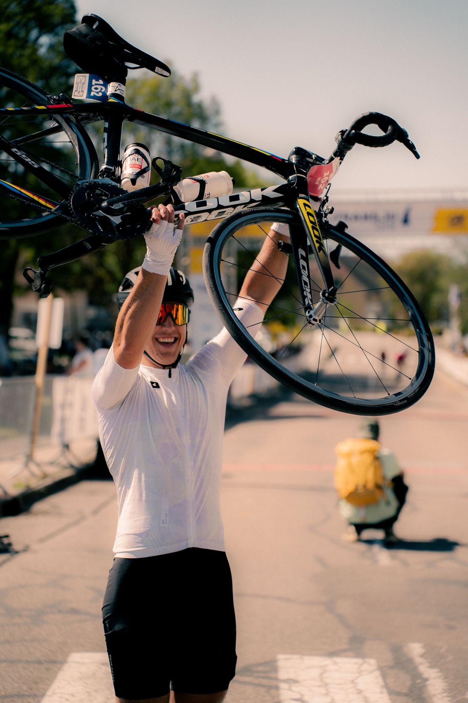 Cyclist in white jersey triumphantly raises black road bike overhead, celebrating victory on sunny urban street.
