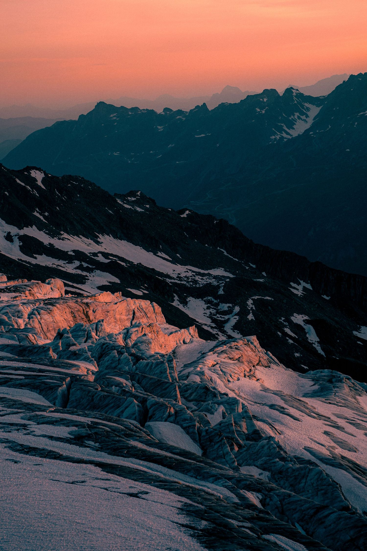 Glacier crevasses glowing orange in dramatic sunset light with layered mountain silhouettes behind.
