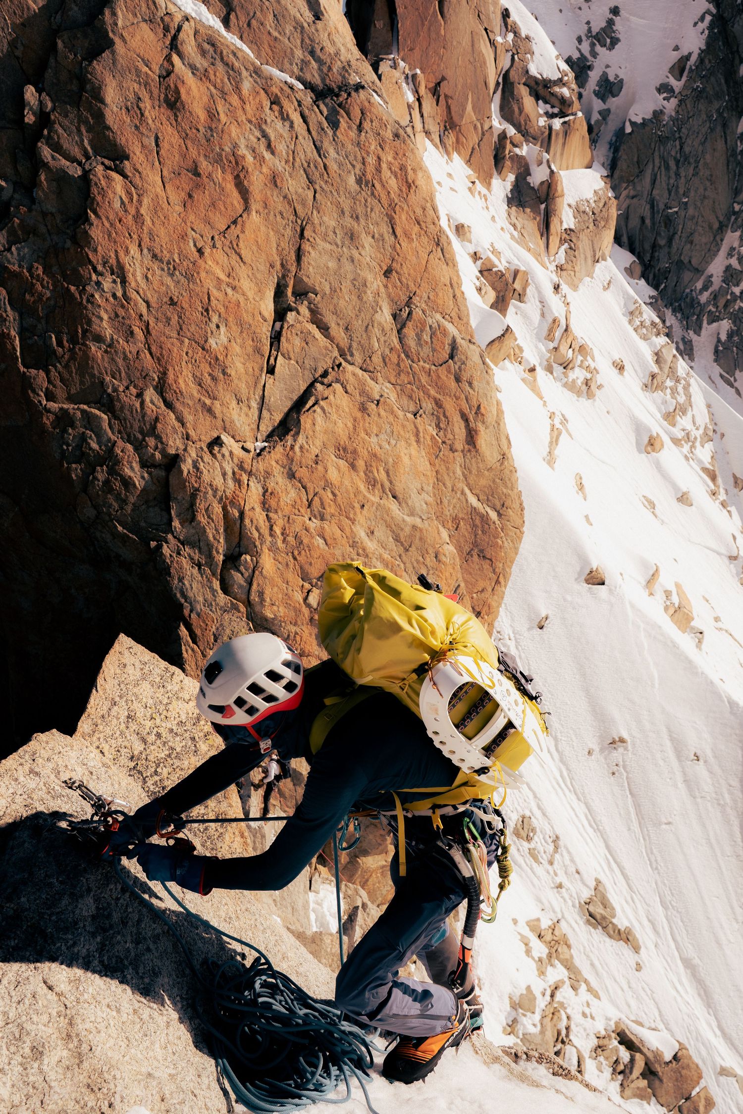 Two climbers in helmets and yellow packs scale a rocky mountain face with snow-covered slopes below.