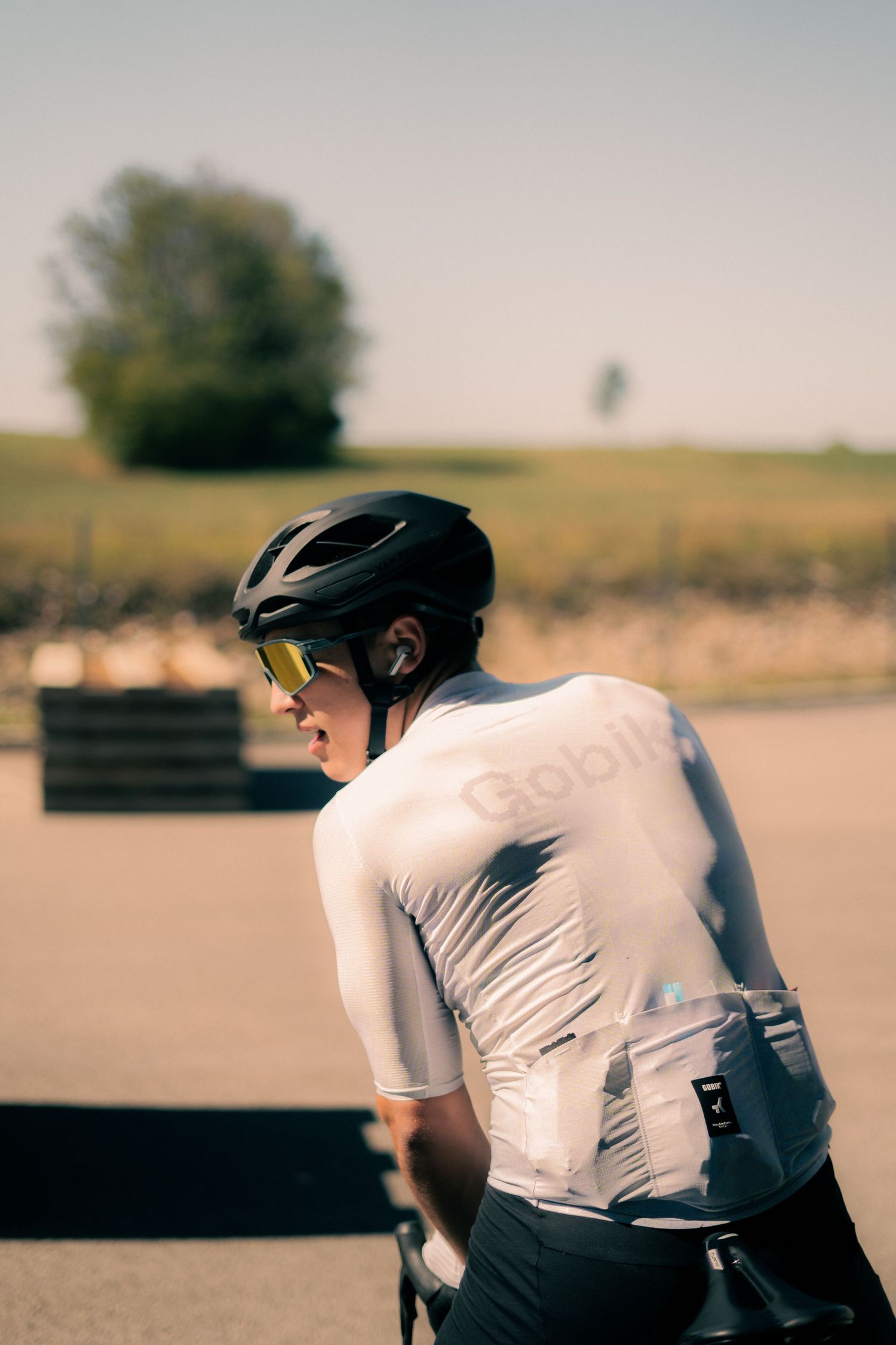 Cyclist in white Gobik jersey, black helmet, and yellow sunglasses posing outdoors on a sunny day.