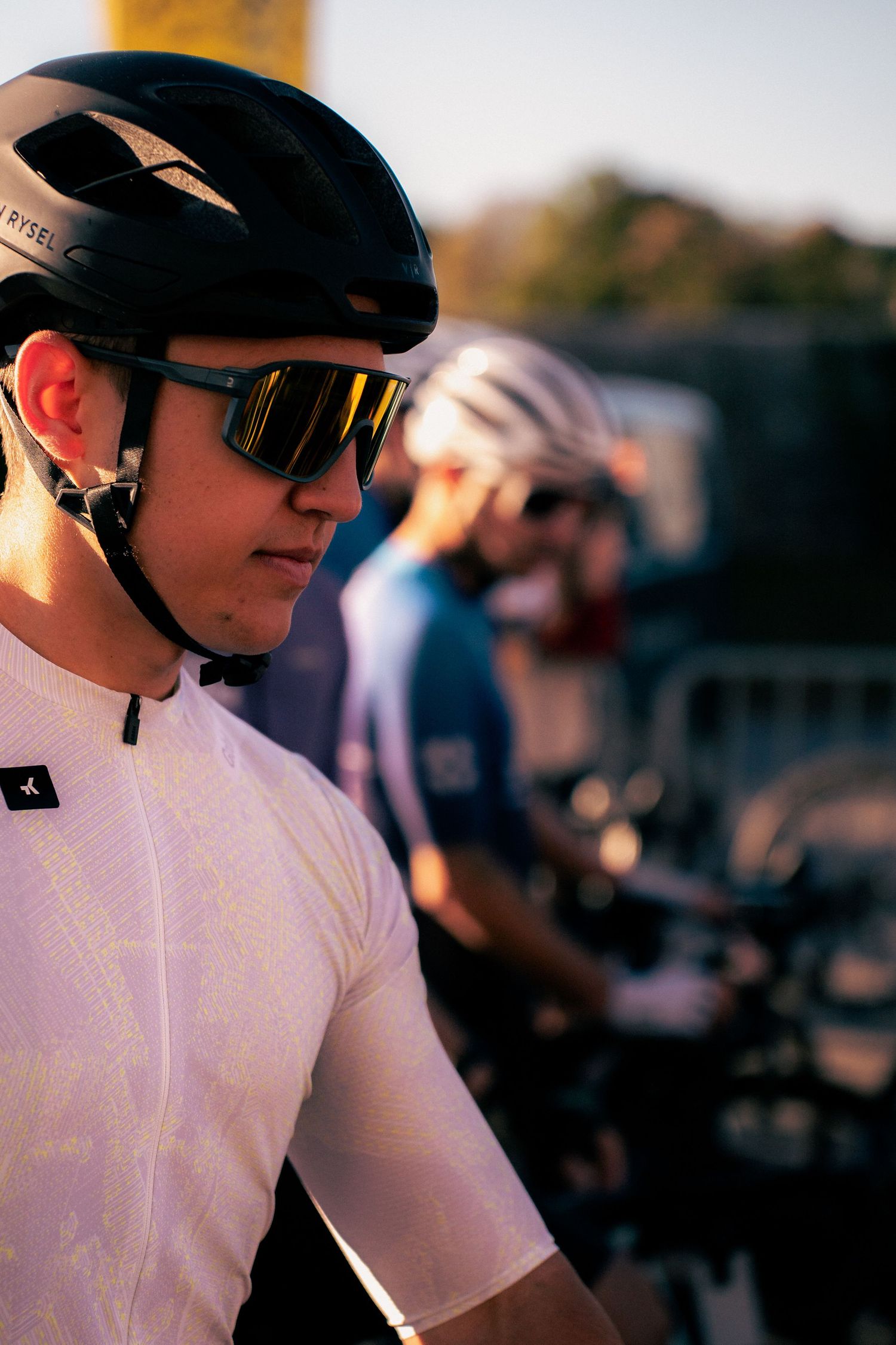 Cyclist in black helmet and yellow sunglasses wearing white jersey at cycling event with riders in background.