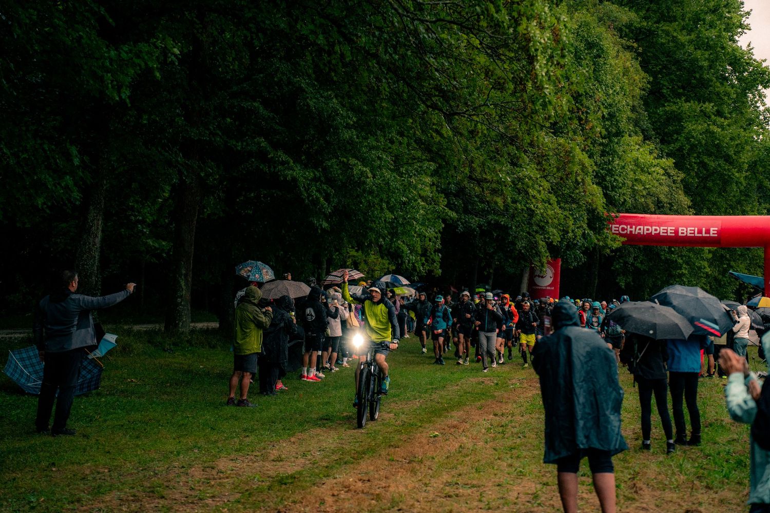 Runners gather under trees at rainy trail race start line at dusk.
