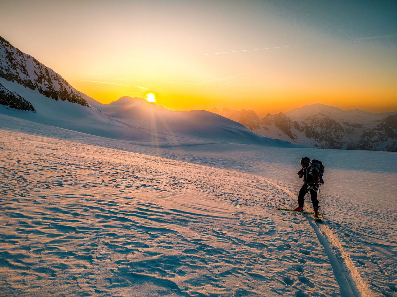 A lone skier ascends a snowy mountain slope at golden sunrise with glowing orange sky and distant peaks.
