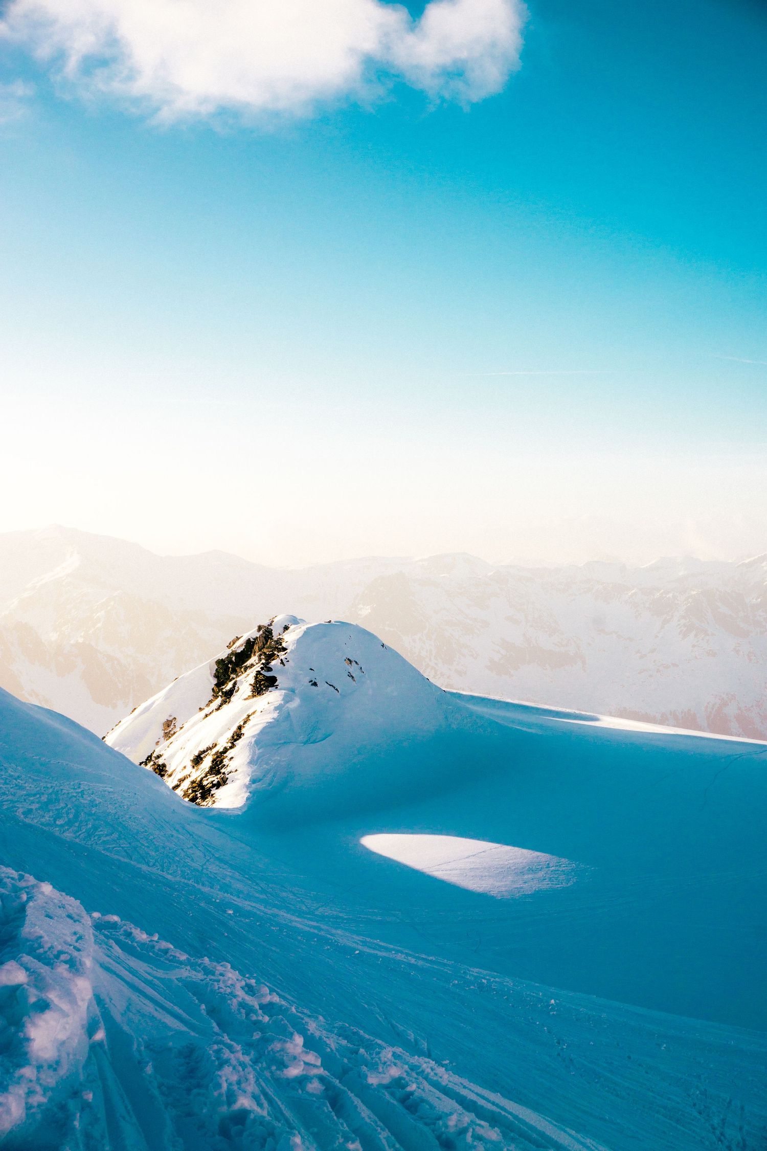 Snow-covered mountain peaks under a clear blue sky with distant alpine ranges bathed in warm sunlight.