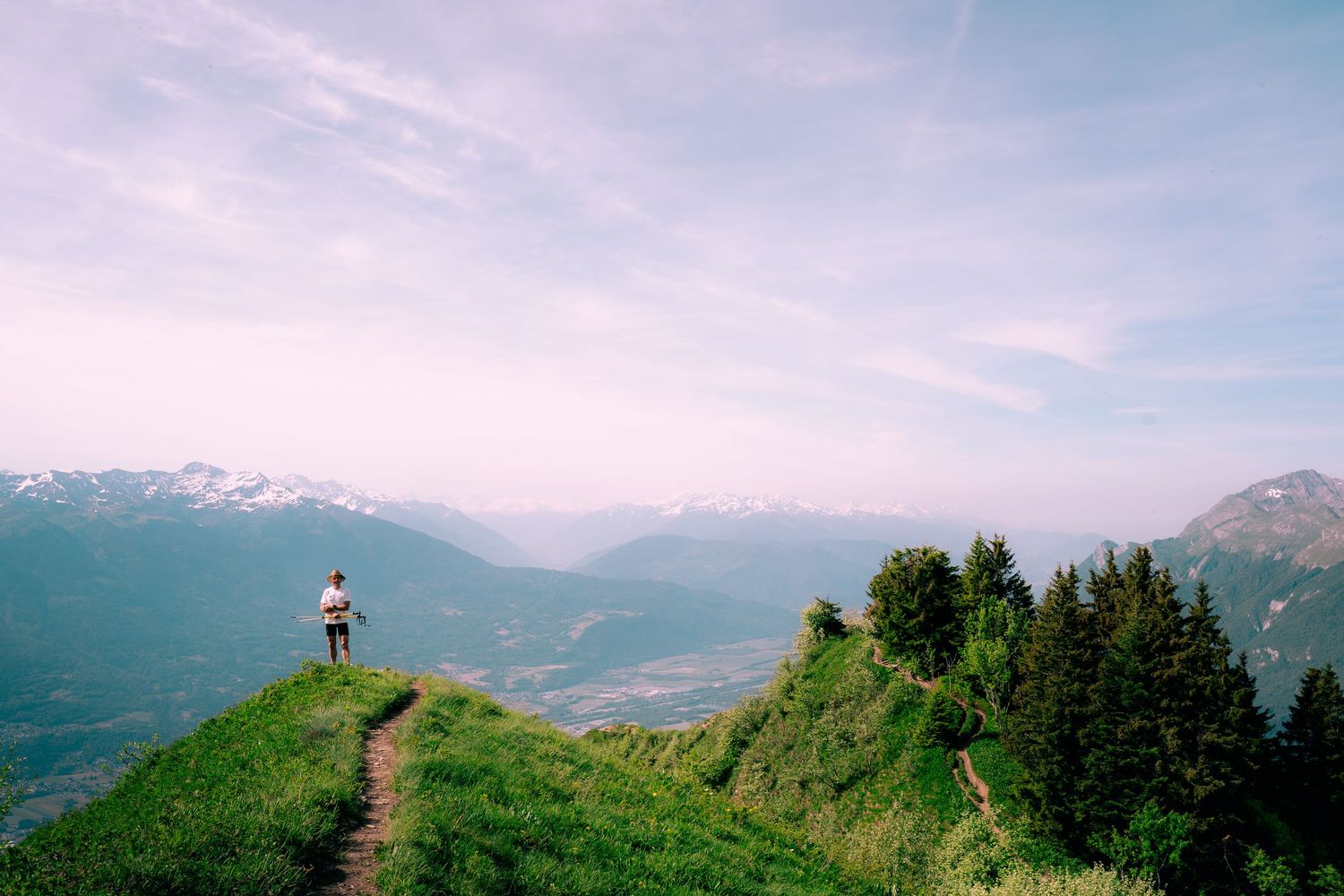 Lone hiker stands on lush green mountain ridge overlooking scenic alpine valley with snow-capped peaks.