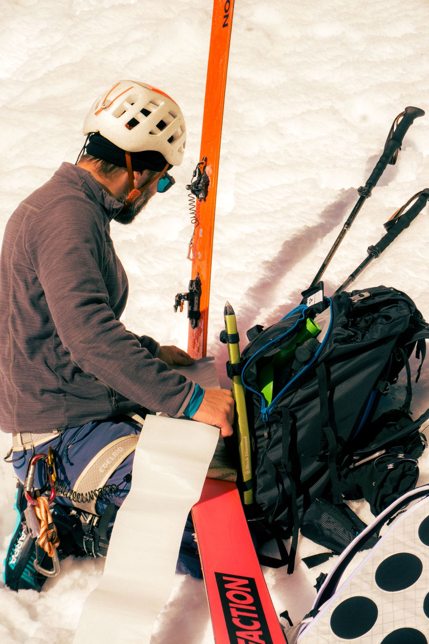 A skier in a white helmet prepares gear on snowy ground with skis, poles, and a backpack.