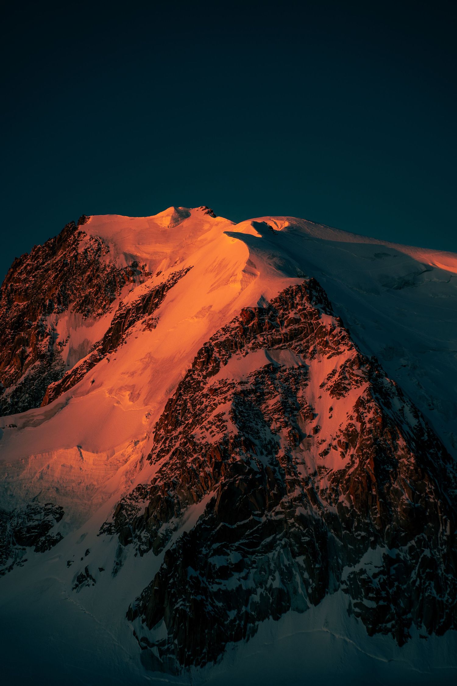 Steep rocky snow-covered mountain peak dramatically lit in fiery orange alpenglow at sunset.