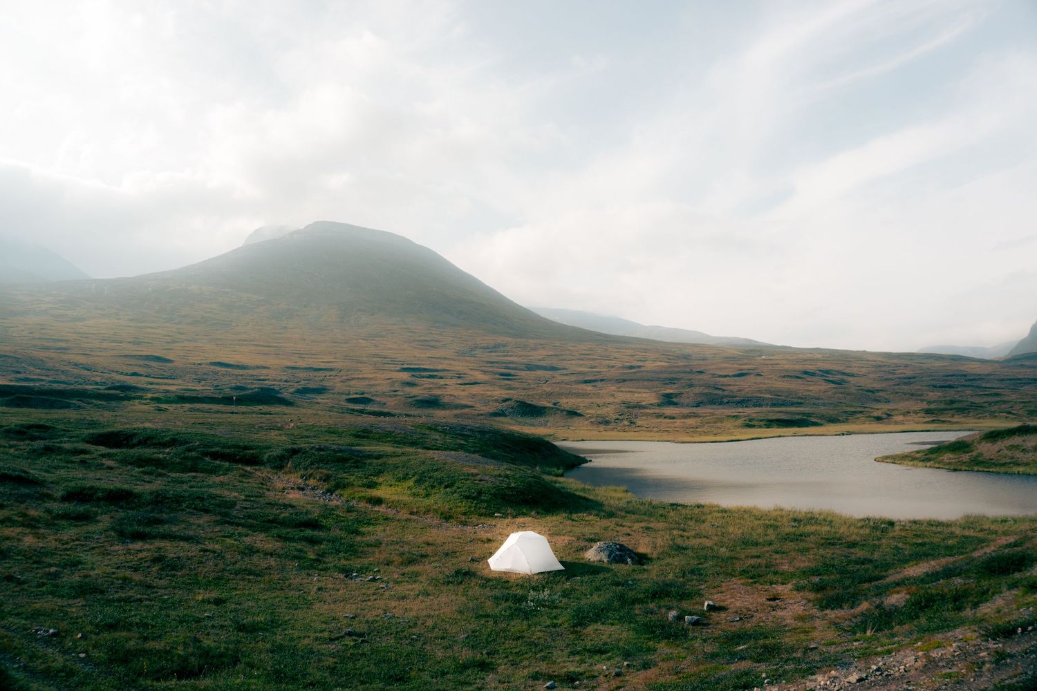 A lone white tent pitched beside a small lake in a vast misty green highland valley landscape.