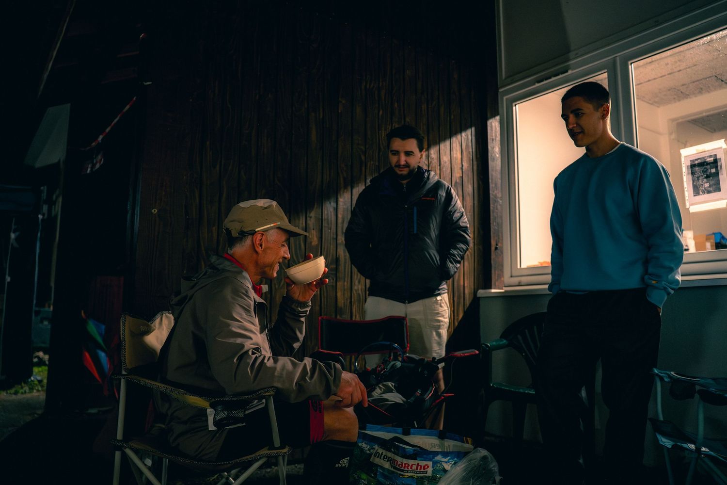 Three men having a nighttime conversation at an outdoor event, one seated in a camping chair under dim lighting.