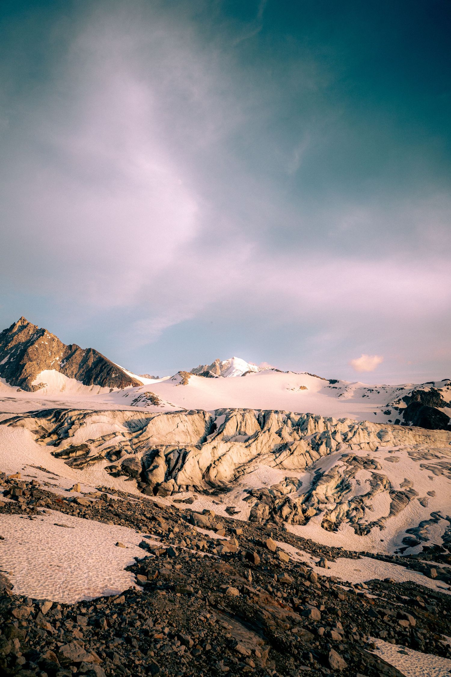 Rugged glacier landscape with crevasses and snow-covered peaks under a cloudy sky at sunset.