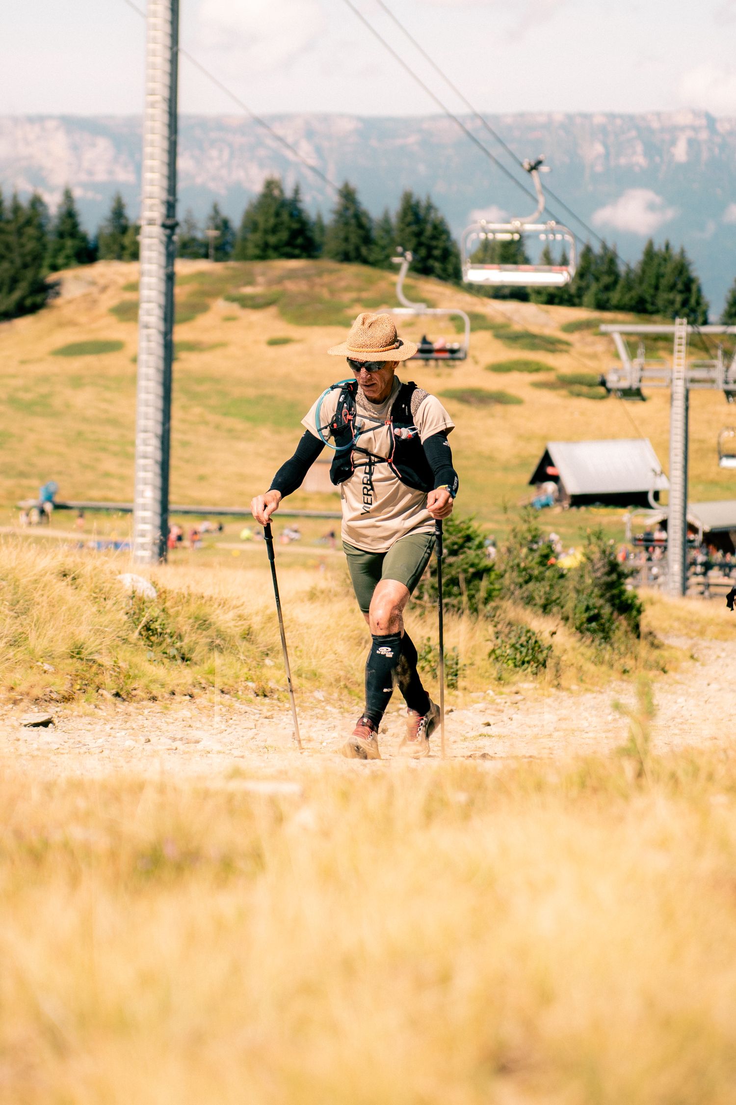 Male hiker with trekking poles and hat walking on a mountain trail near a ski lift in summer.