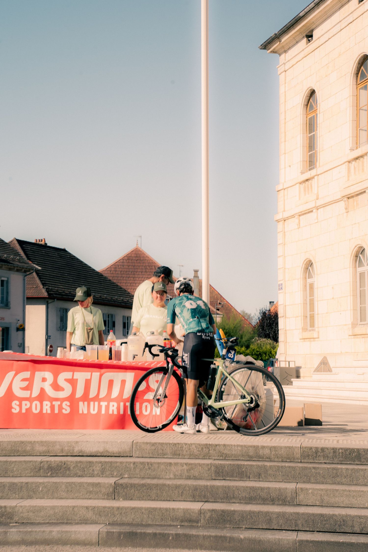 Cyclists gather near an Overstim Sports Nutrition banner at a cycling event outside a historic stone building.
