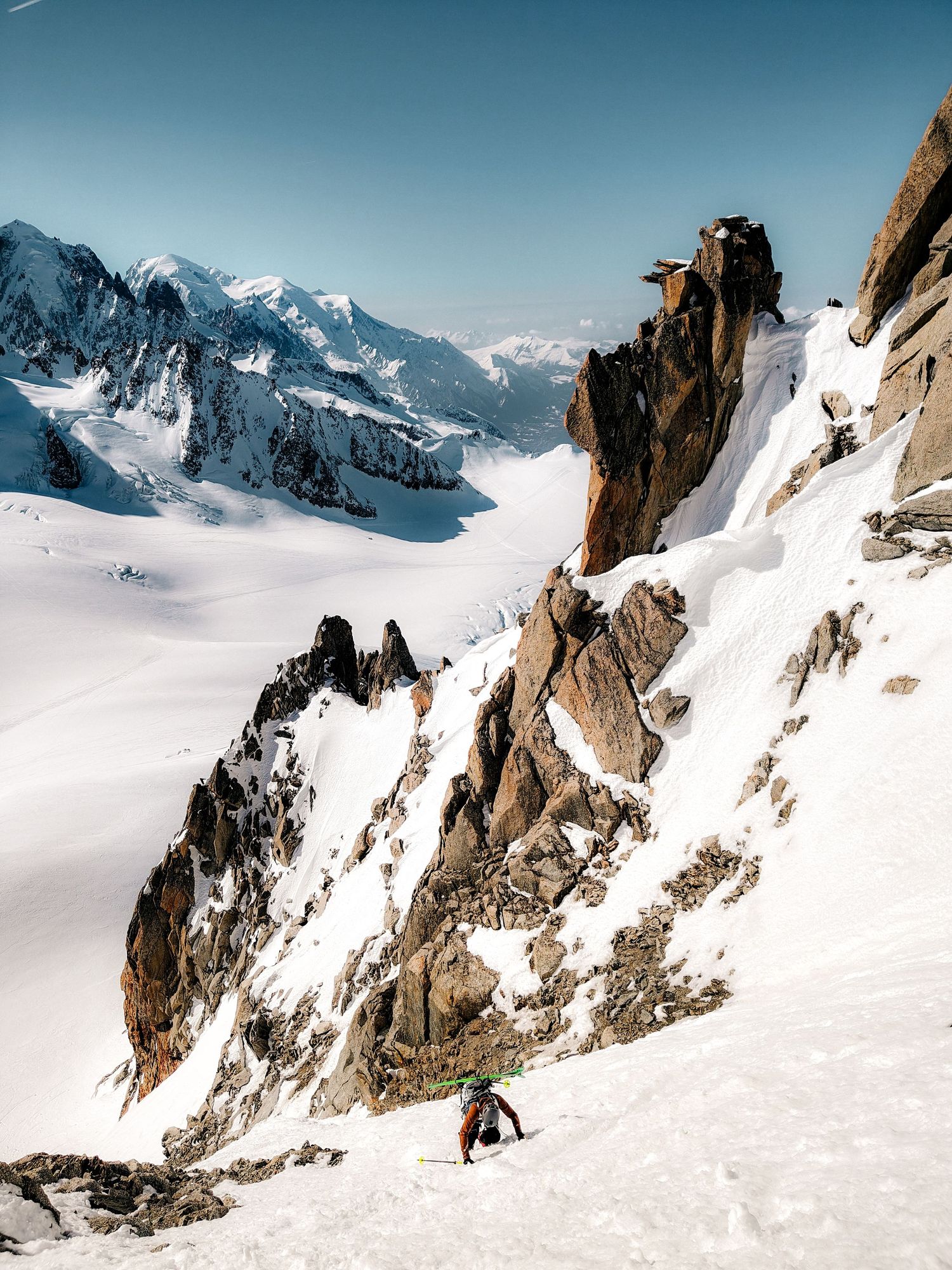 A lone skier descends a steep snow-covered alpine slope surrounded by rocky peaks and vast mountain glacier below.