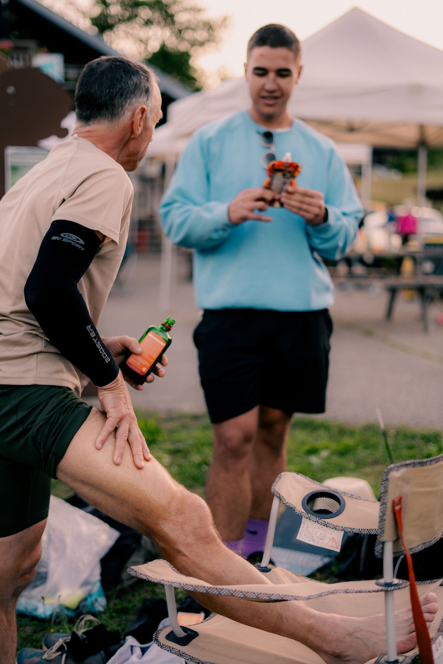 Male athlete stretching his leg at an outdoor race aid station while a young man stands nearby.