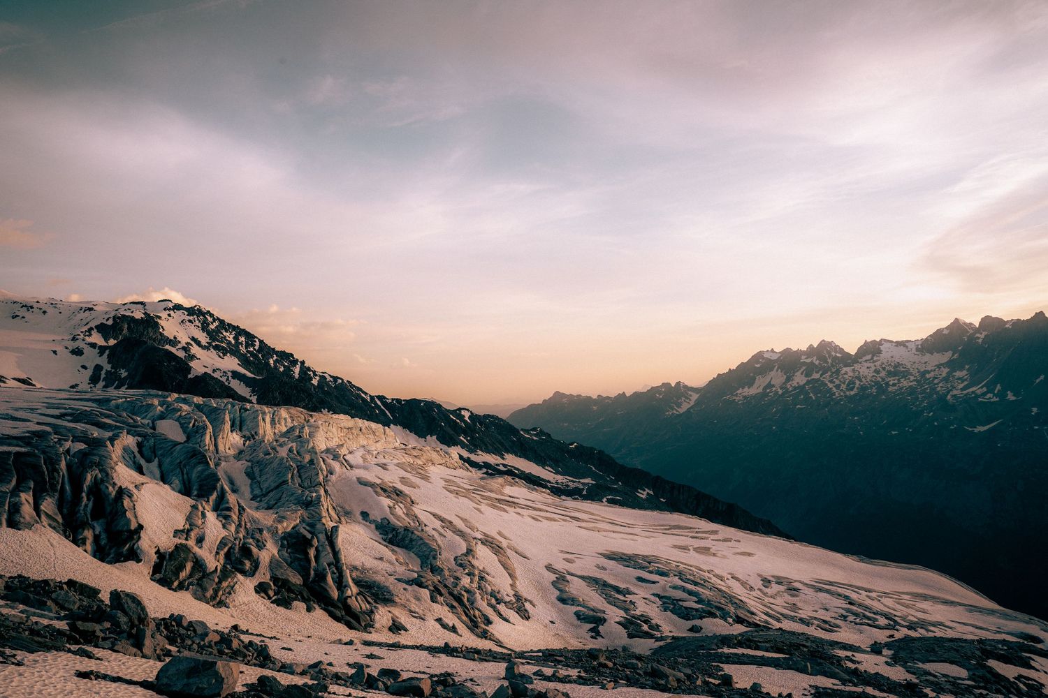 Expansive snow-covered mountain range at sunset with soft pink and purple hues in the sky.