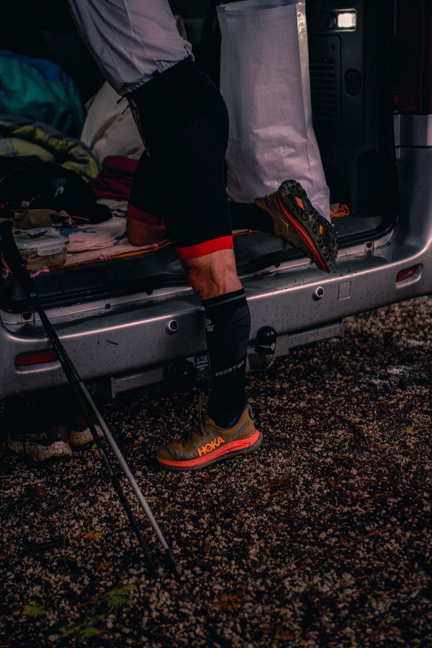 Trail runner wearing Hoka shoes and compression socks prepares gear at vehicle tailgate before race.