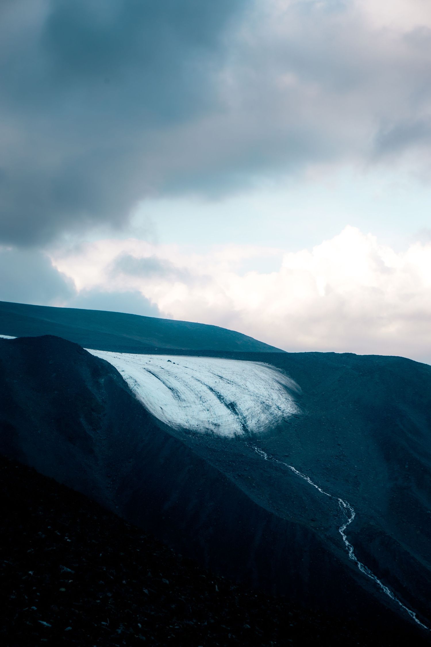 Snow-capped mountain glacier with meltwater stream flowing down rocky slopes under dramatic clouds.