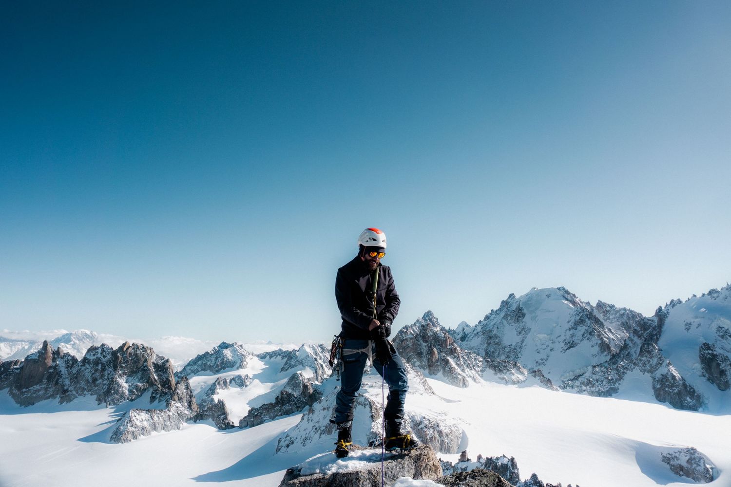 A mountaineer stands triumphantly on a snowy peak wearing a helmet and goggles with vast alpine ranges behind.