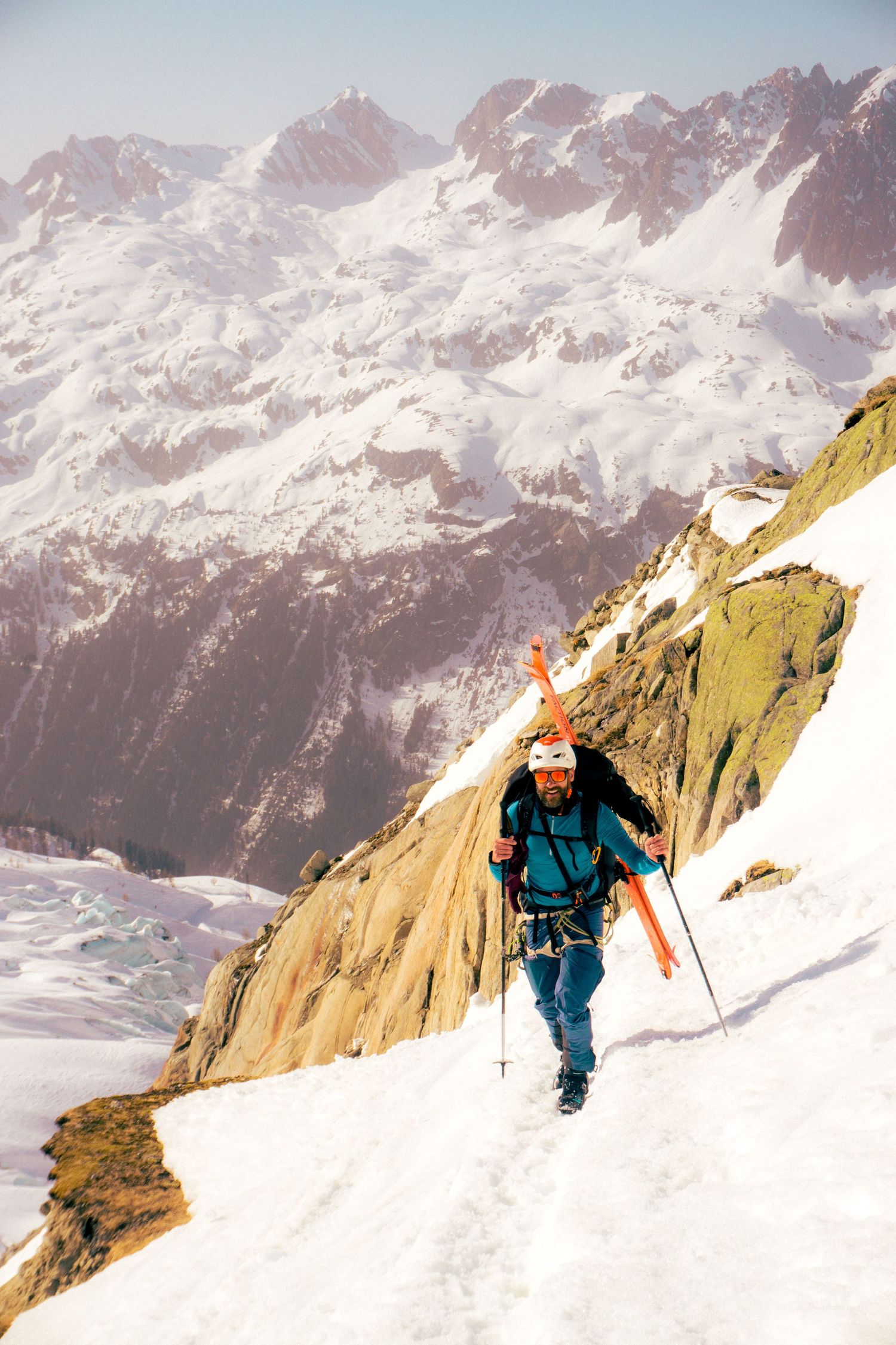 A ski mountaineer climbs a snowy alpine ridge with skis on back and poles, surrounded by dramatic snow-capped mountain peaks.