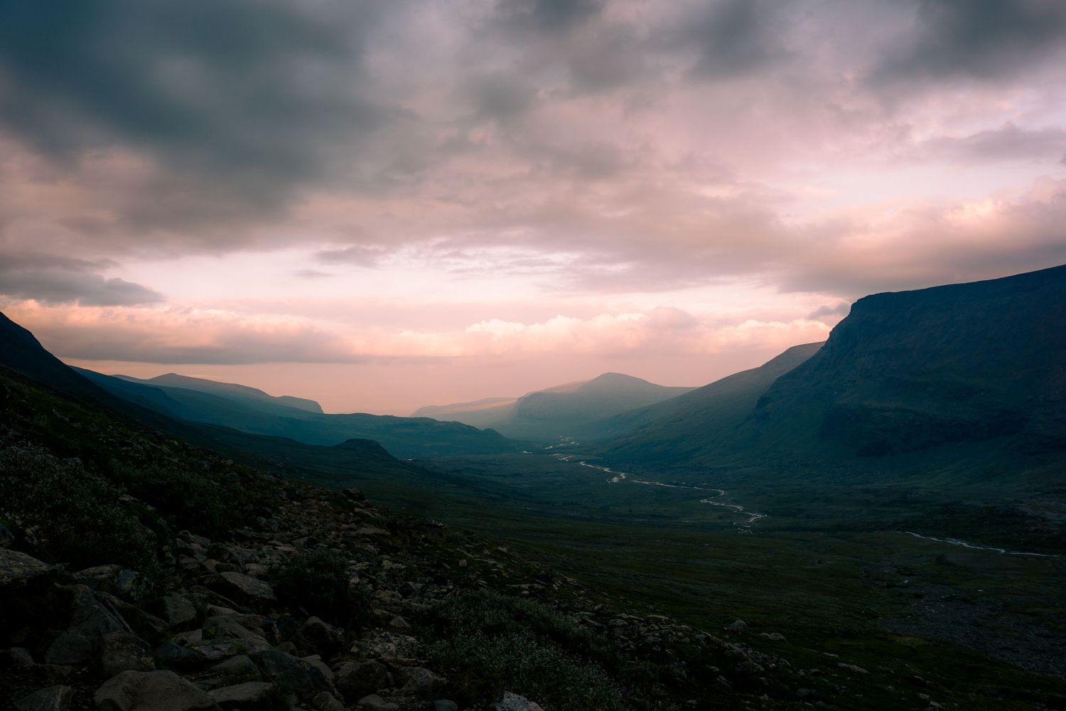 Moody twilight mountain valley landscape with rocky foreground and dramatic pink and purple clouds.