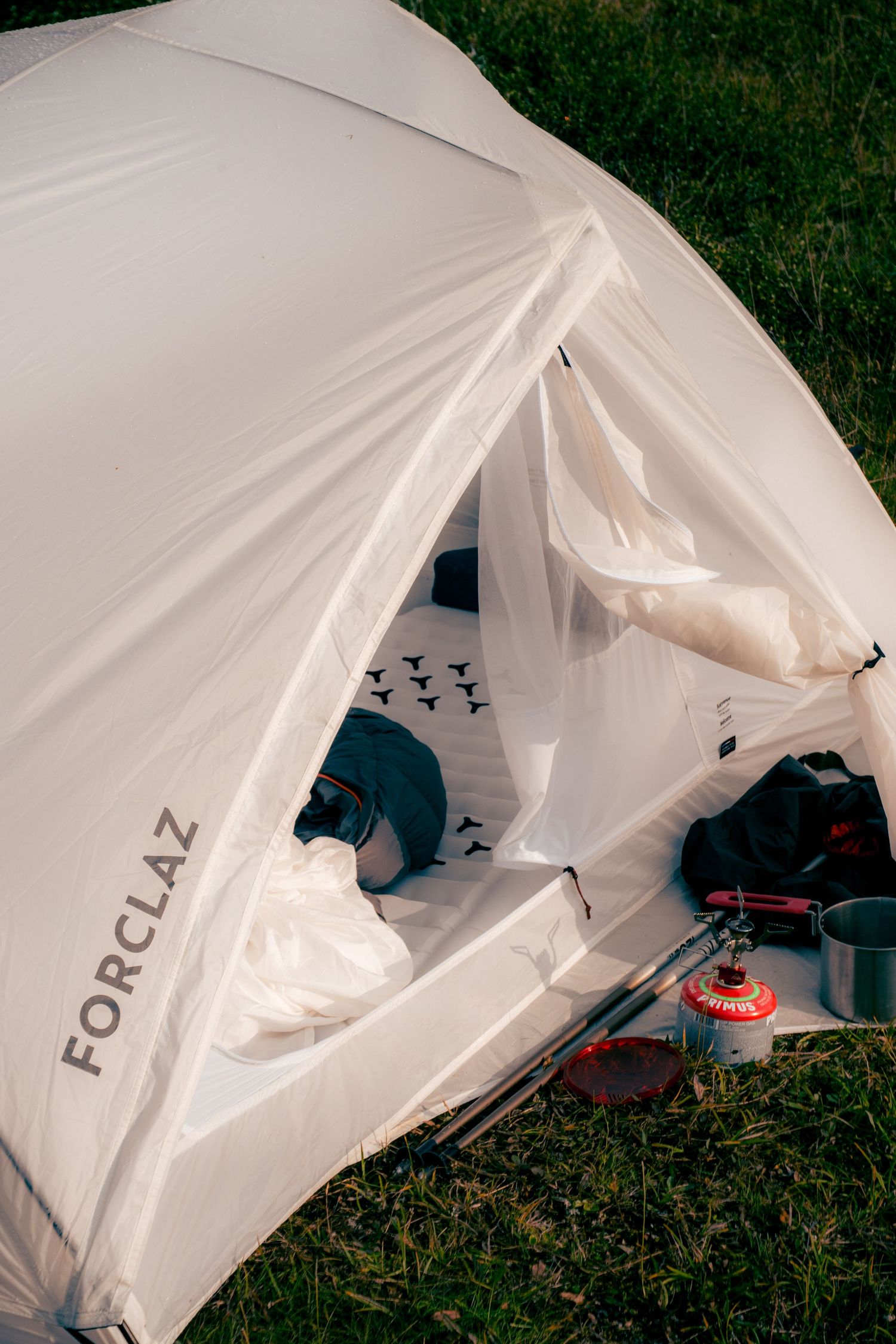 Close-up of open Forclaz brand white camping tent with cooking stove and gear on green grass.