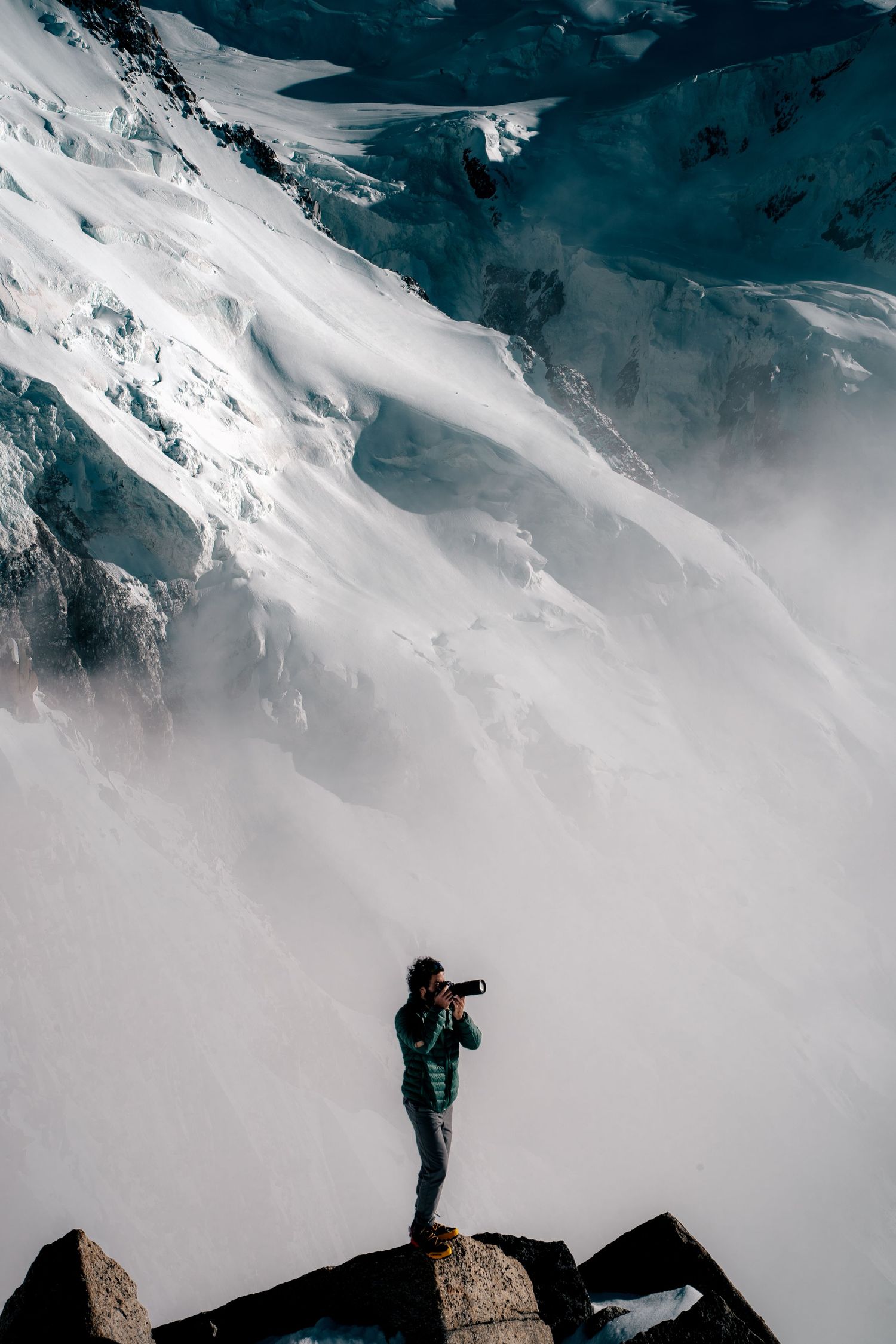 A photographer in a green jacket captures sweeping views of glacial snow slopes from a rocky mountain perch.
