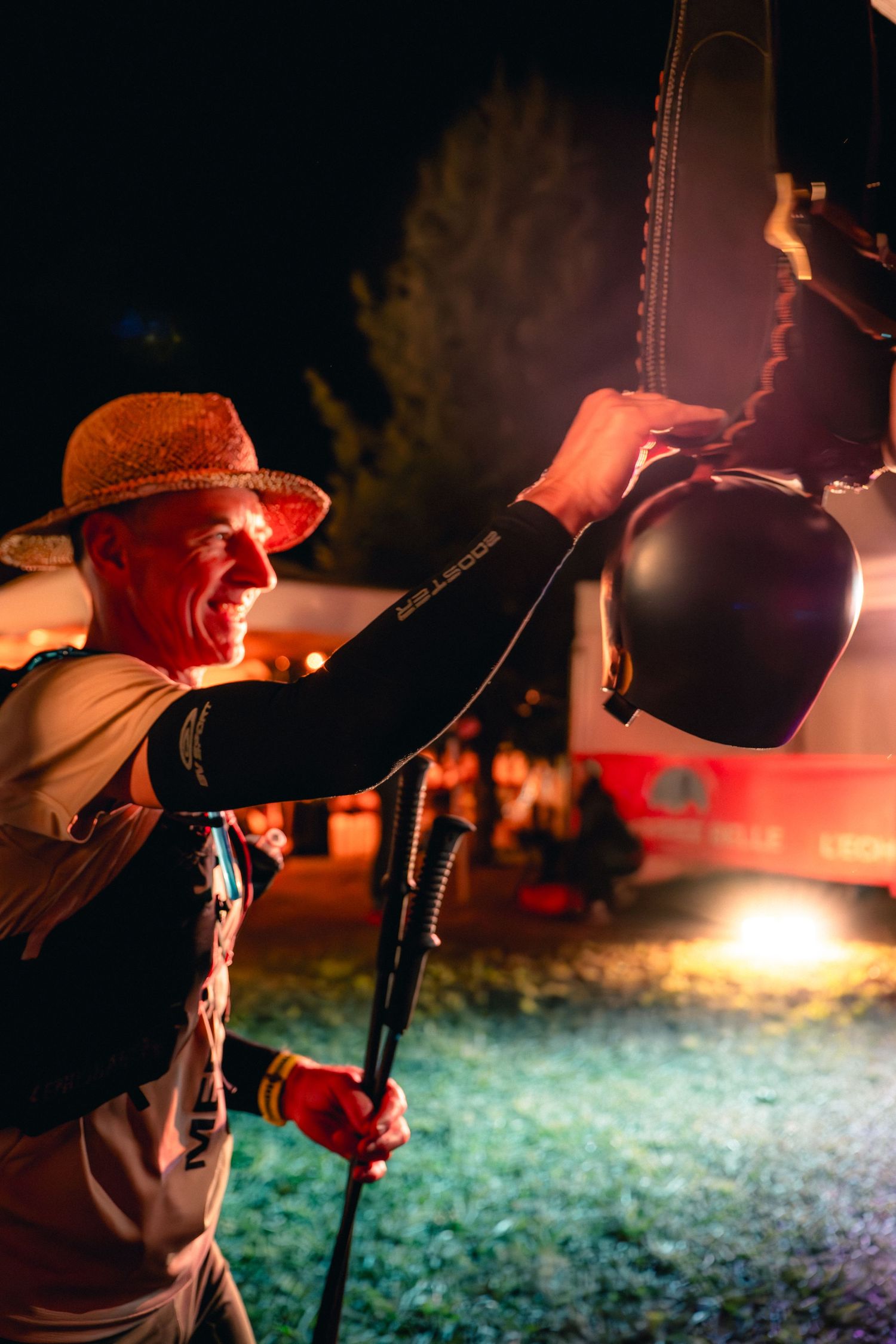 Smiling male trail runner holding a helmet at a night race checkpoint with warm lighting.