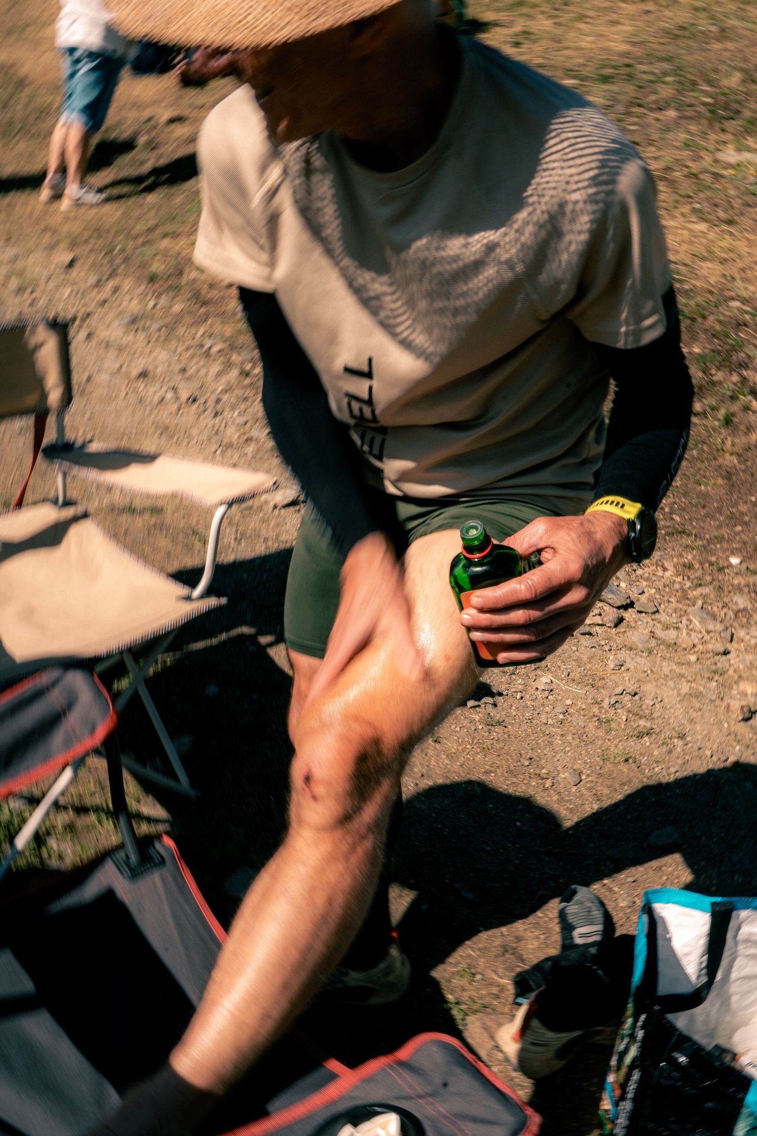 Trail runner applying recovery oil to knee while resting in chair at outdoor race checkpoint.